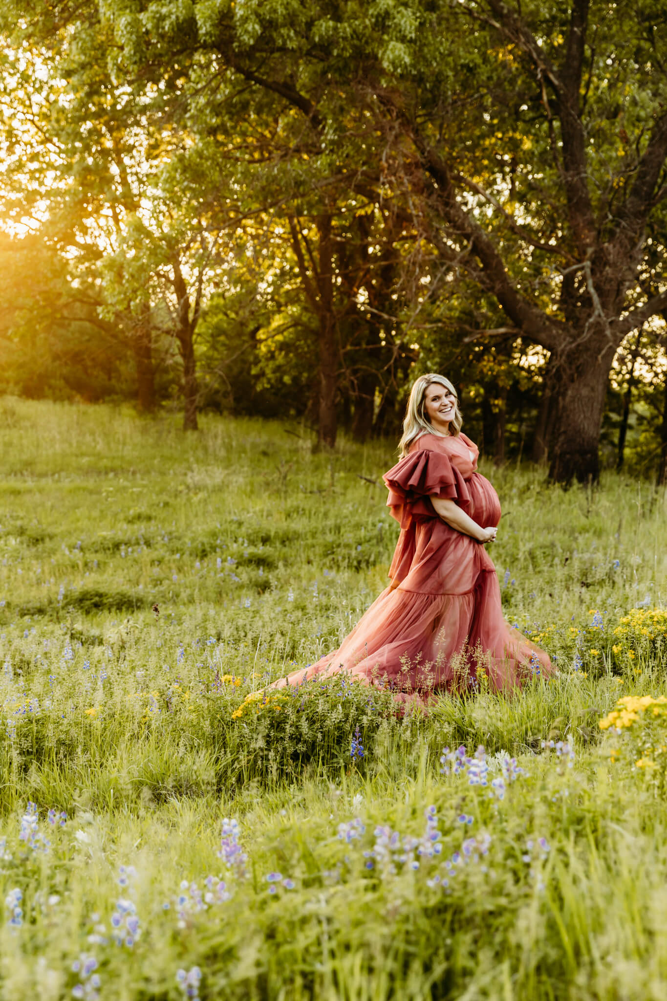 expecting mama in a long robe with ruffled shoulders dancing in a flower field and smiling as the sunshines through for blog about Froedtert Birth Center