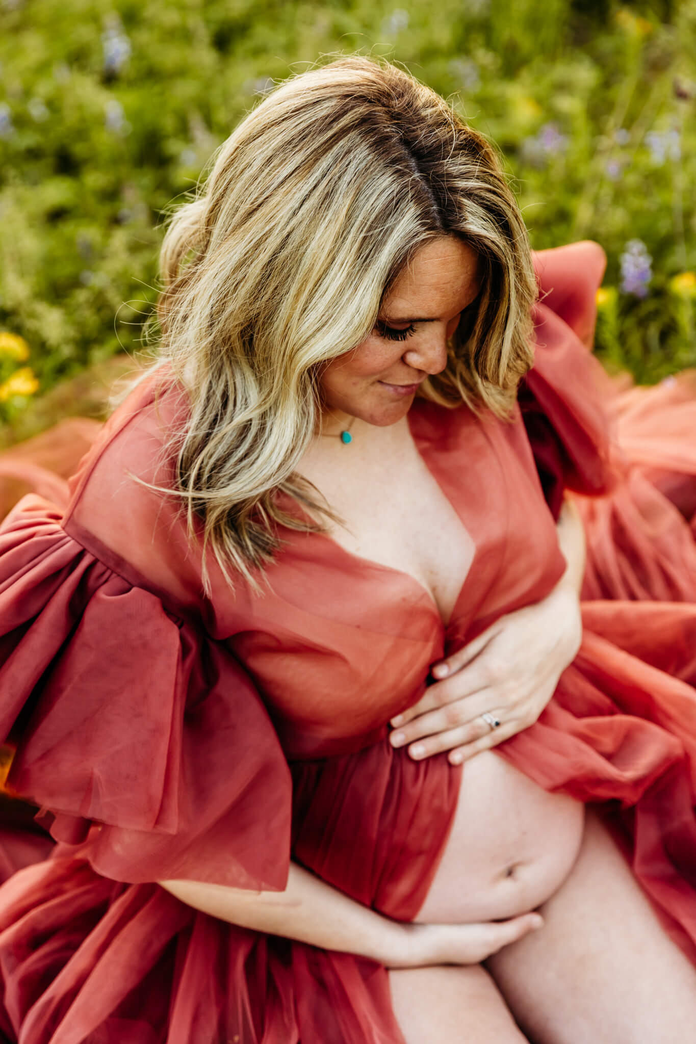 expecting mama kneeling in a flower field in a flowy robe and holding her bare baby bump while looking down