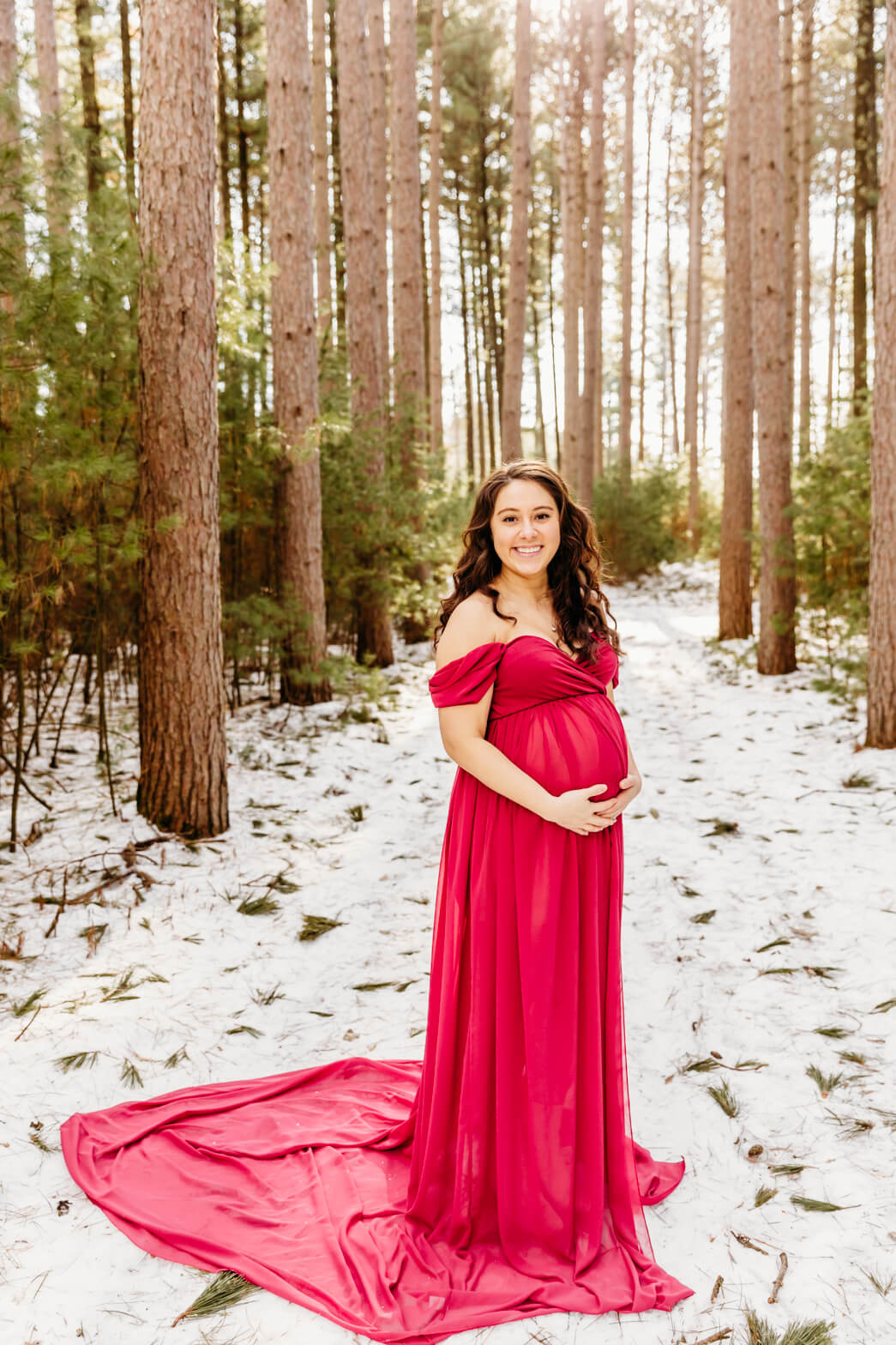 A mom to be in a long flowing maternity gown holds her bump with standing in a snow covered path through a pine forest