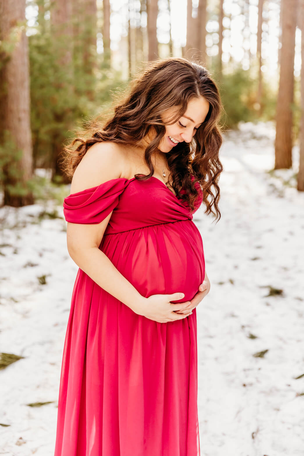 A mom to be looks down at her bump while standing in a snowy pine forest froedtert birth center