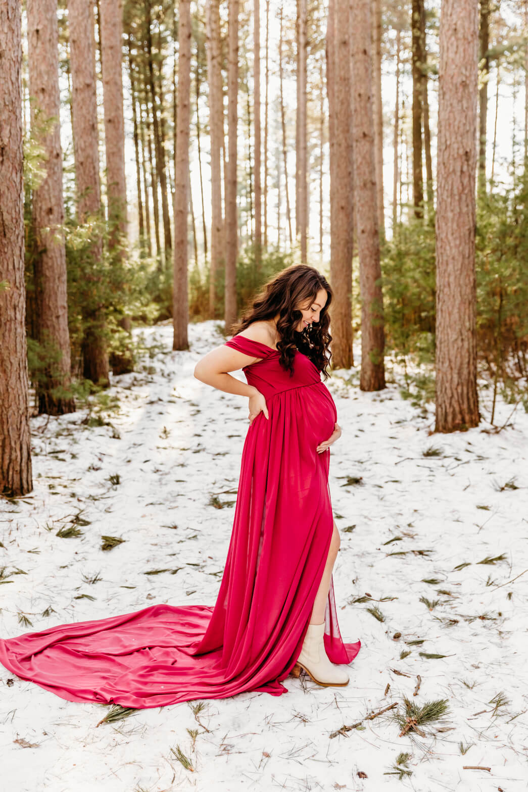 A mom to be in white boots and a pink maternity gown stands in a snowy pine forest froedtert birth center