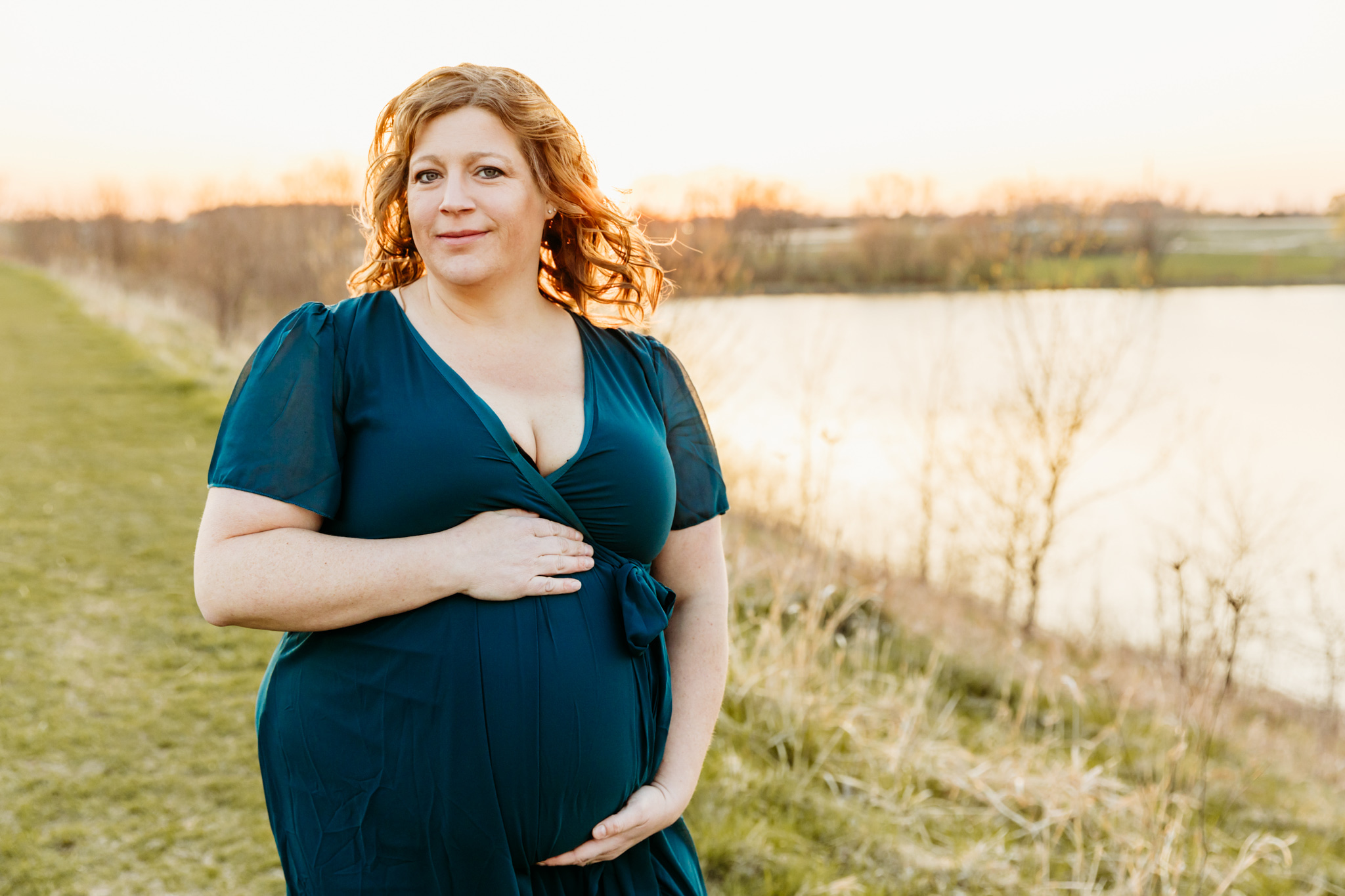 A mom to be stands by a lake in a grassy path while holding her bump in a blue maternity dress