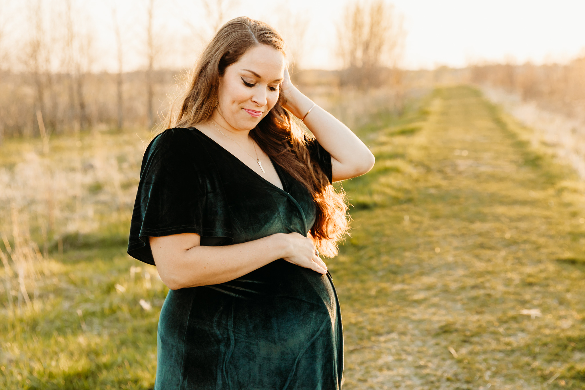 A mother to be looks down at her bump in a green velvet dress while standing in a grassy park path