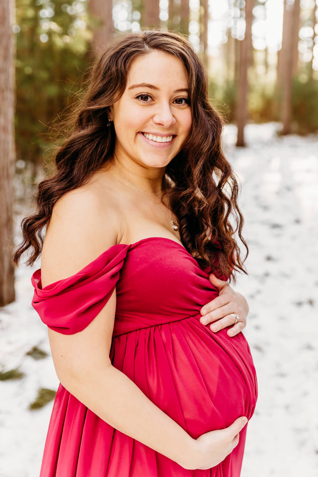 A mother to be holds her bump in a pink maternity gown while standing in a snowy forest