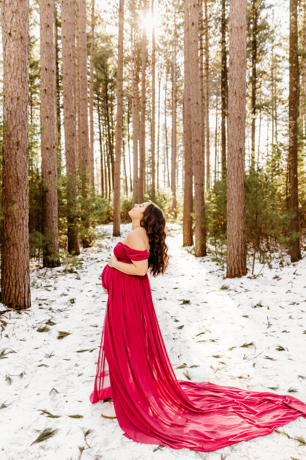 A pregnant woman stands in a snowy forest wearing a long red maternity gown jill coulter