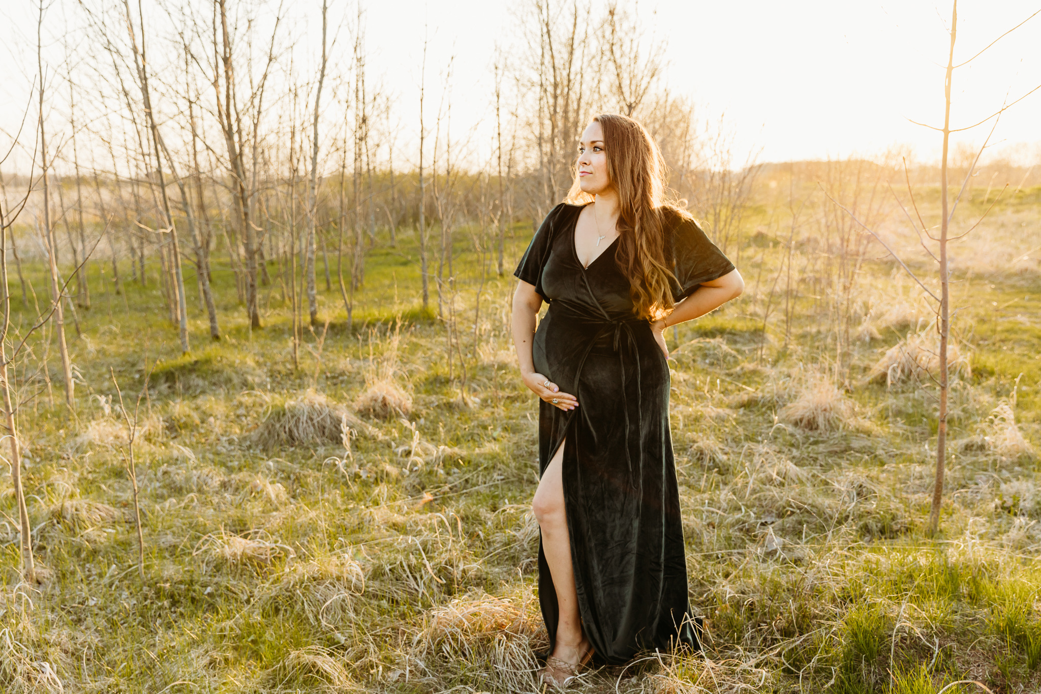 A mom to be holds her bump while standing in a field surrounded by trees with no leaves