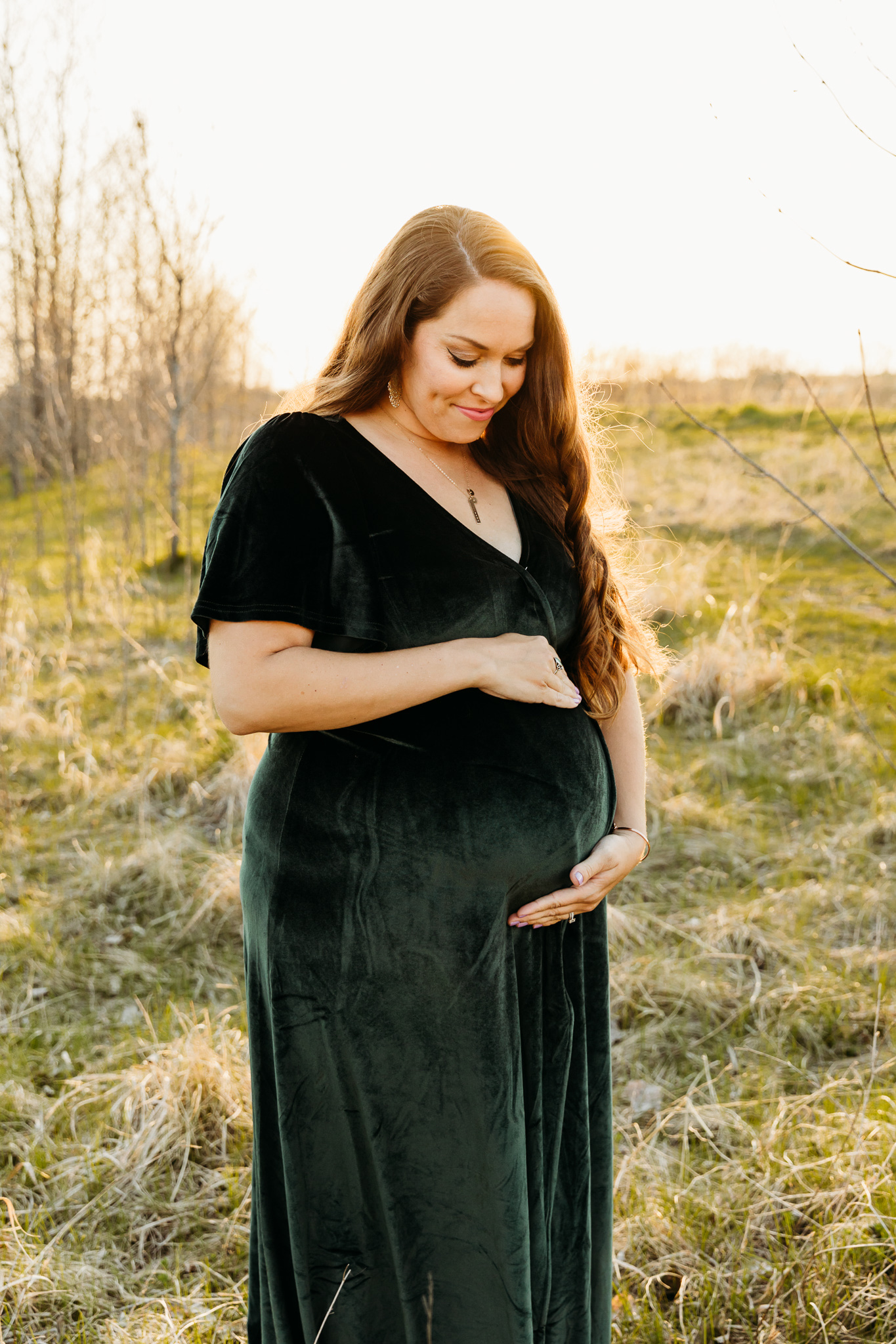 A mother to be stands in a field wearing a green maternity dress while looking down at the bump her hands are holding Milwaukee yoga studios