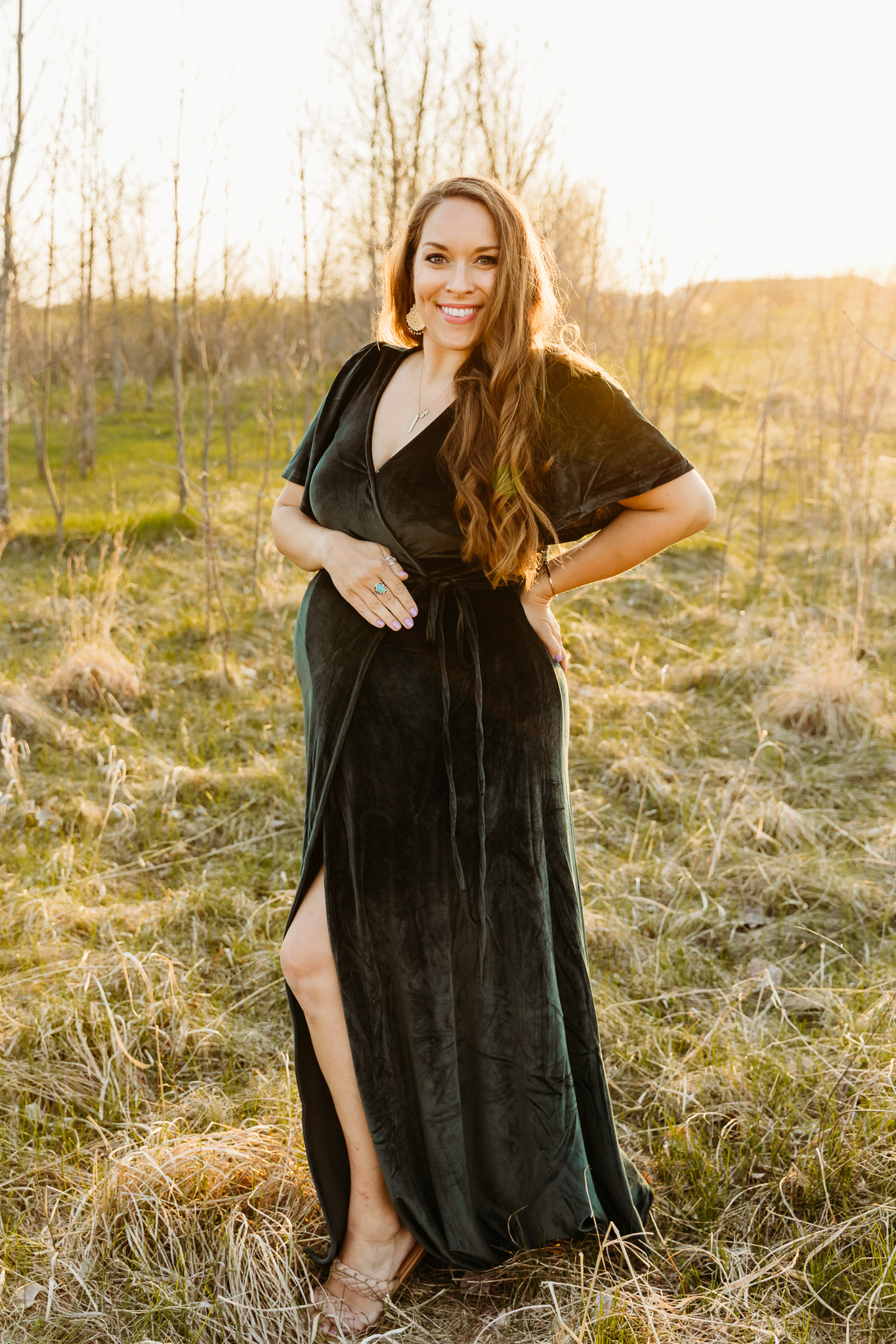 A mother to be in a green velvet maternity dress stands in a field with a hand on her back Milwaukee yoga studios