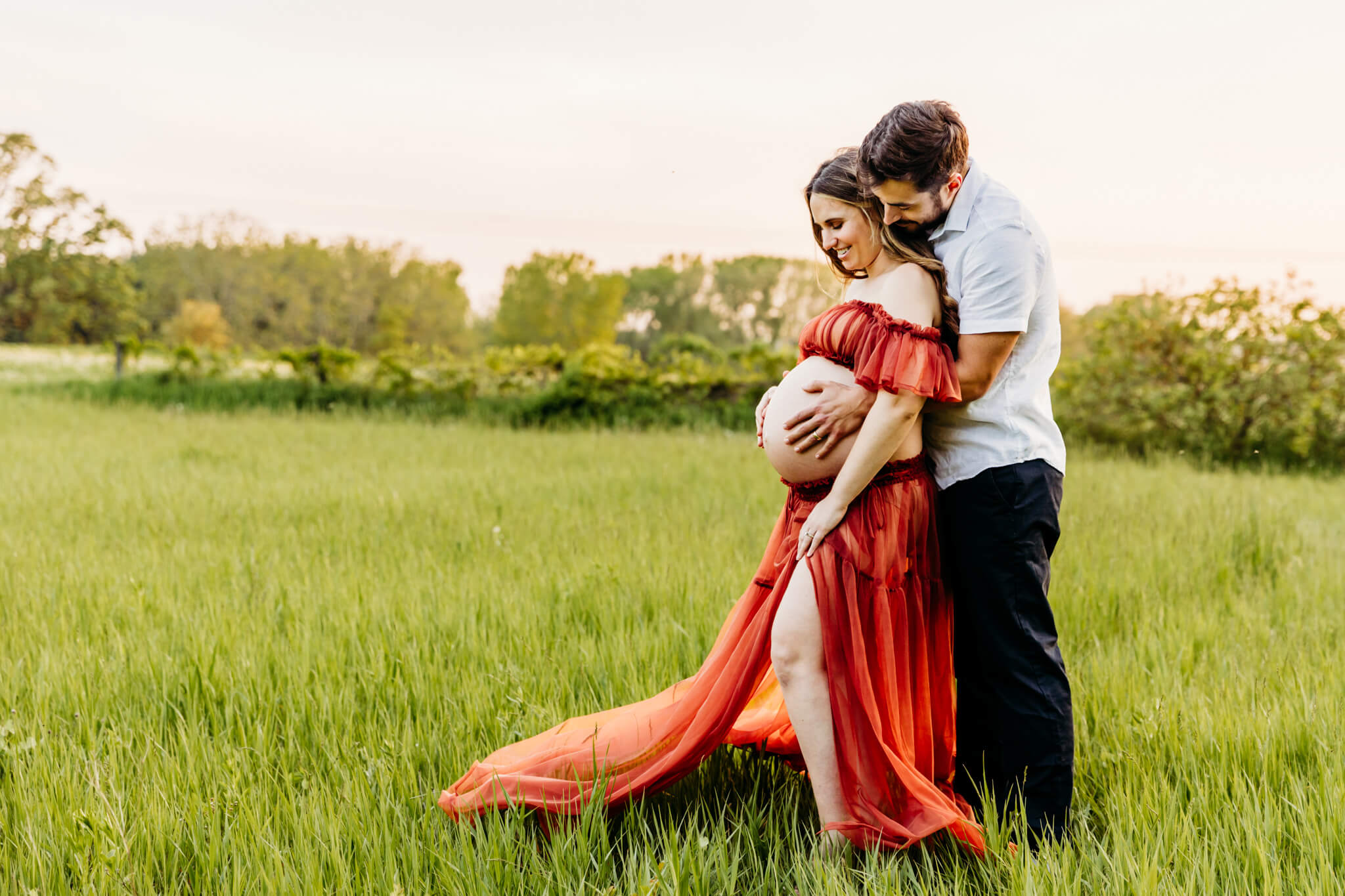 husband holding his wife and wrapping arms around her baby bump as they laugh together in a grassy field for blog about Mothering the Mother