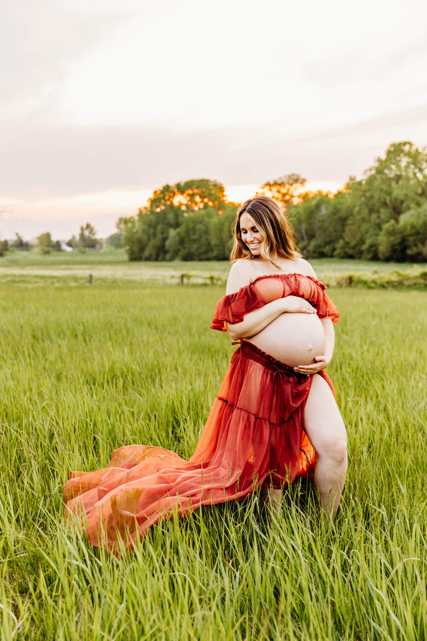 mom to be in orange two piece dress holding her bare baby bump and looking down as sun sets behind her