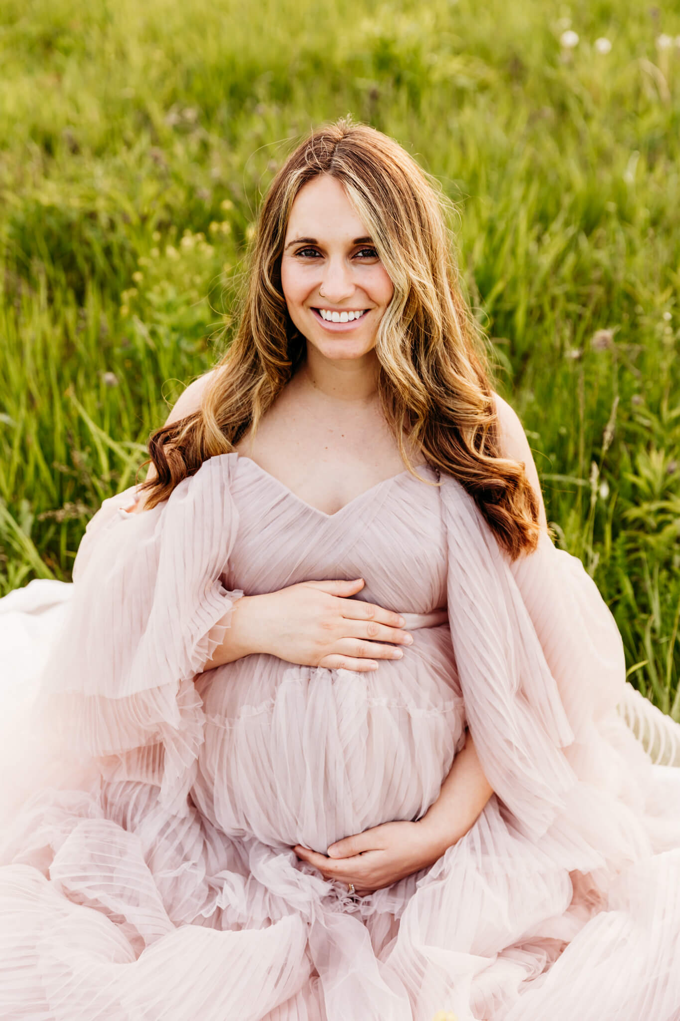 expecting mom in a blush gown holding her baby bump and looking up as she sits in a grassy wildflower field for blog about New Beginnings Pregnancy Center