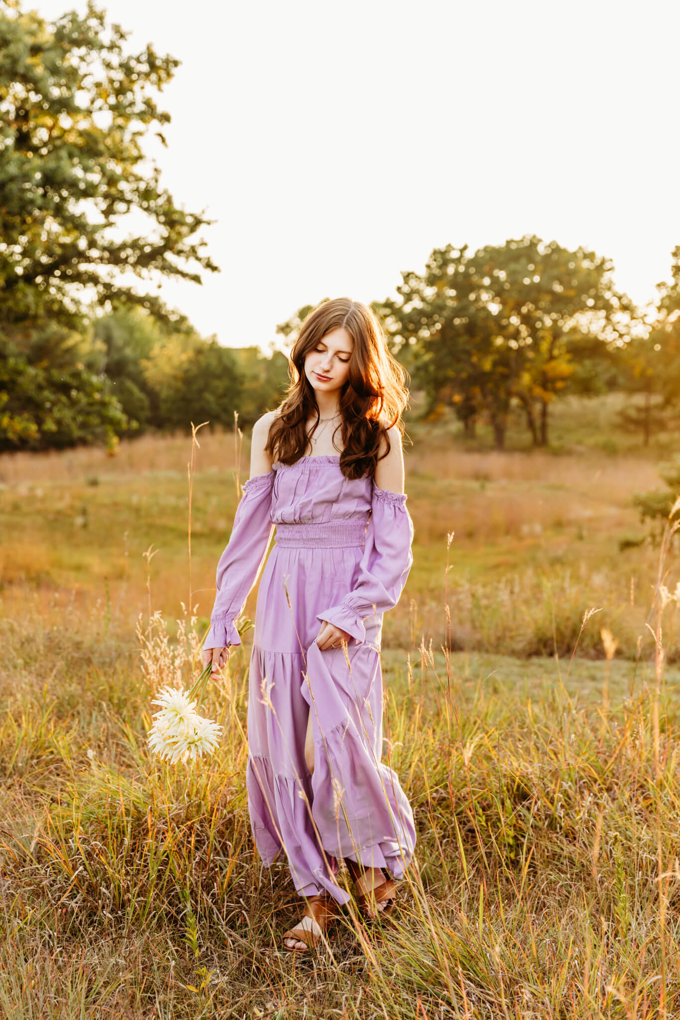 gorgeous teen girl in a flowy purple dress holding flowers and walking in a field at sunset