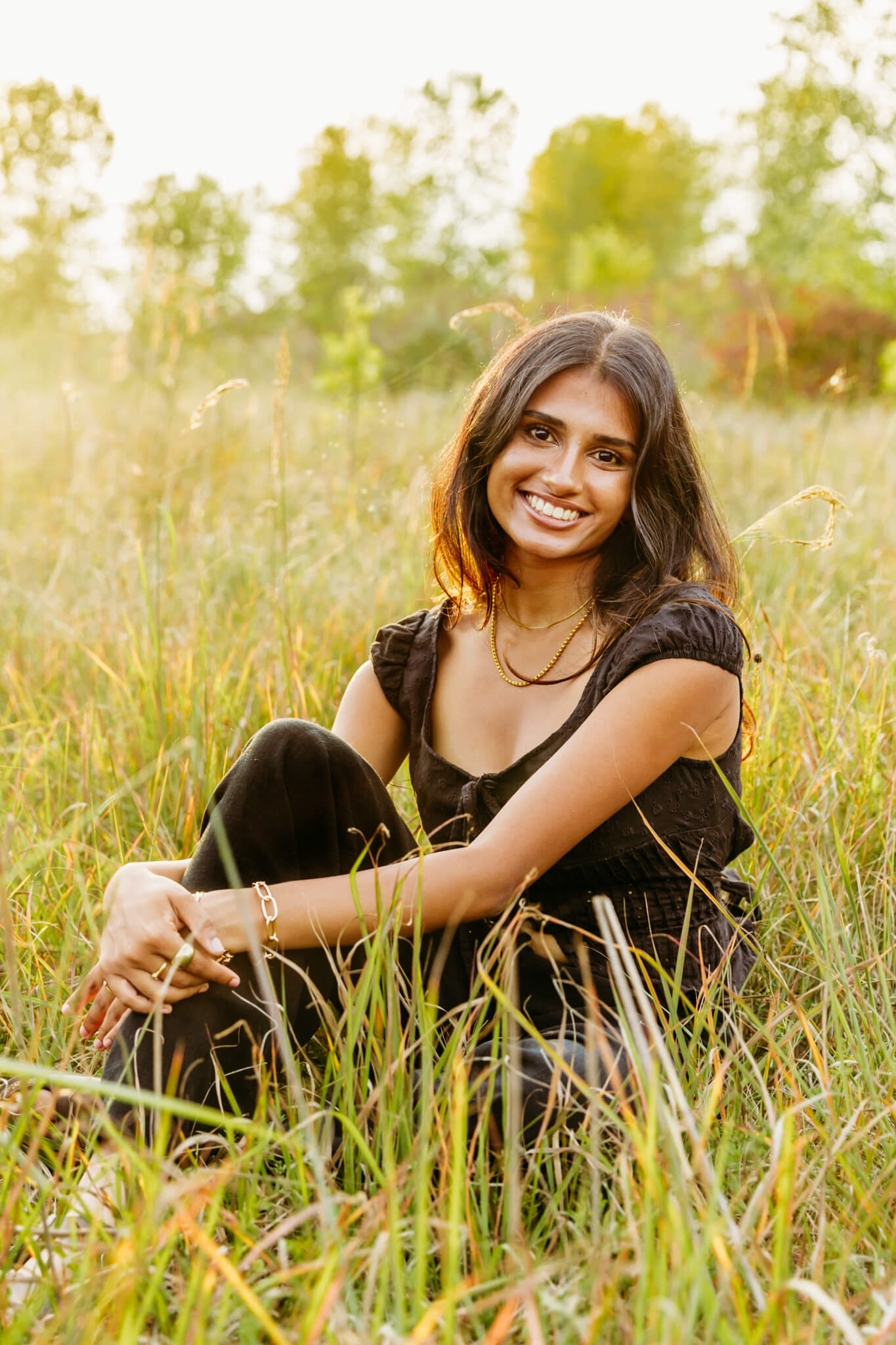 gorgeous high school senior girl from Bay City Christian School in a black jumpsuit sitting in grass at sunset