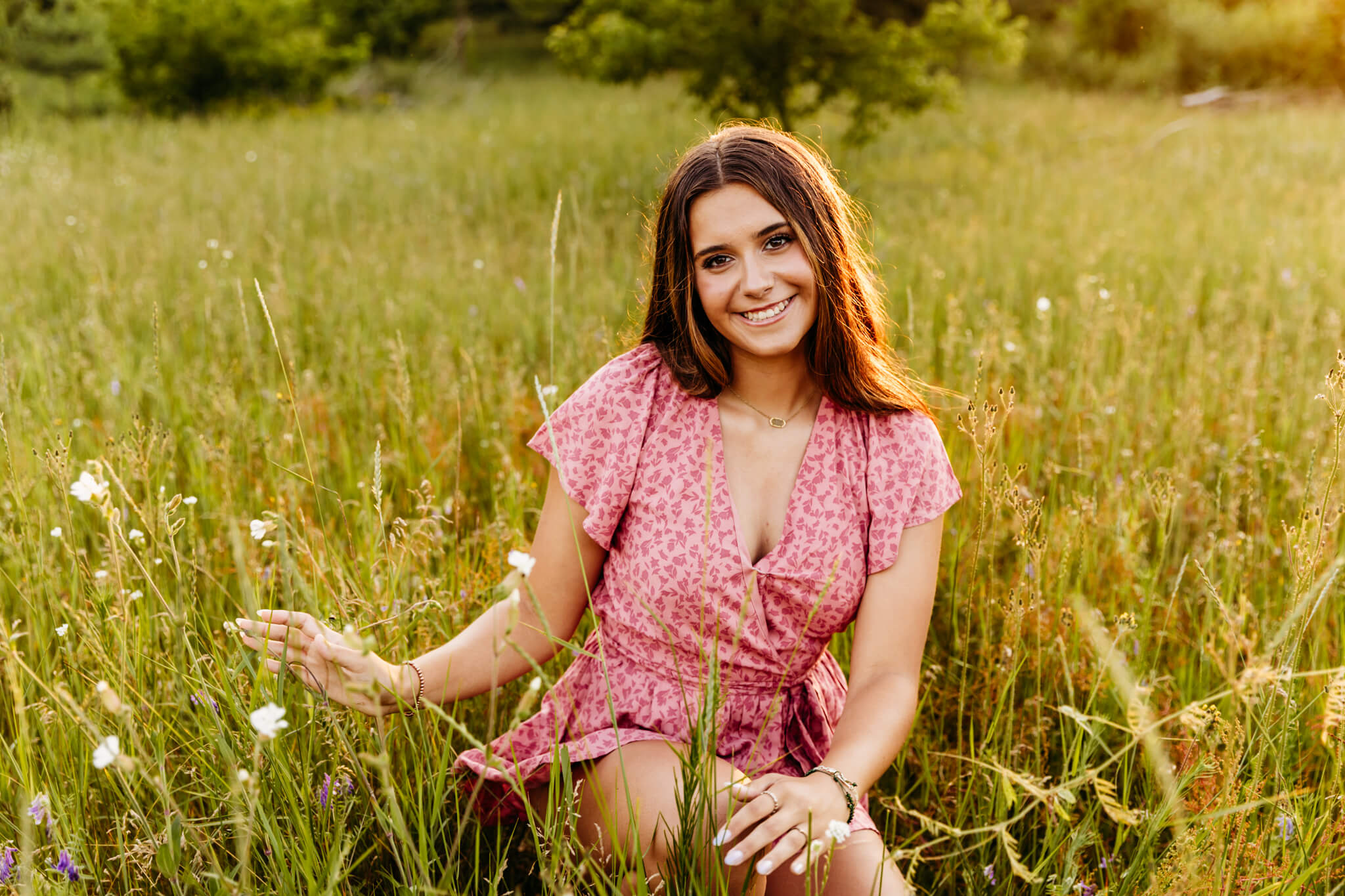 stunning brown haired teen girl in a pink romper kneeling in a grassy field at sunset