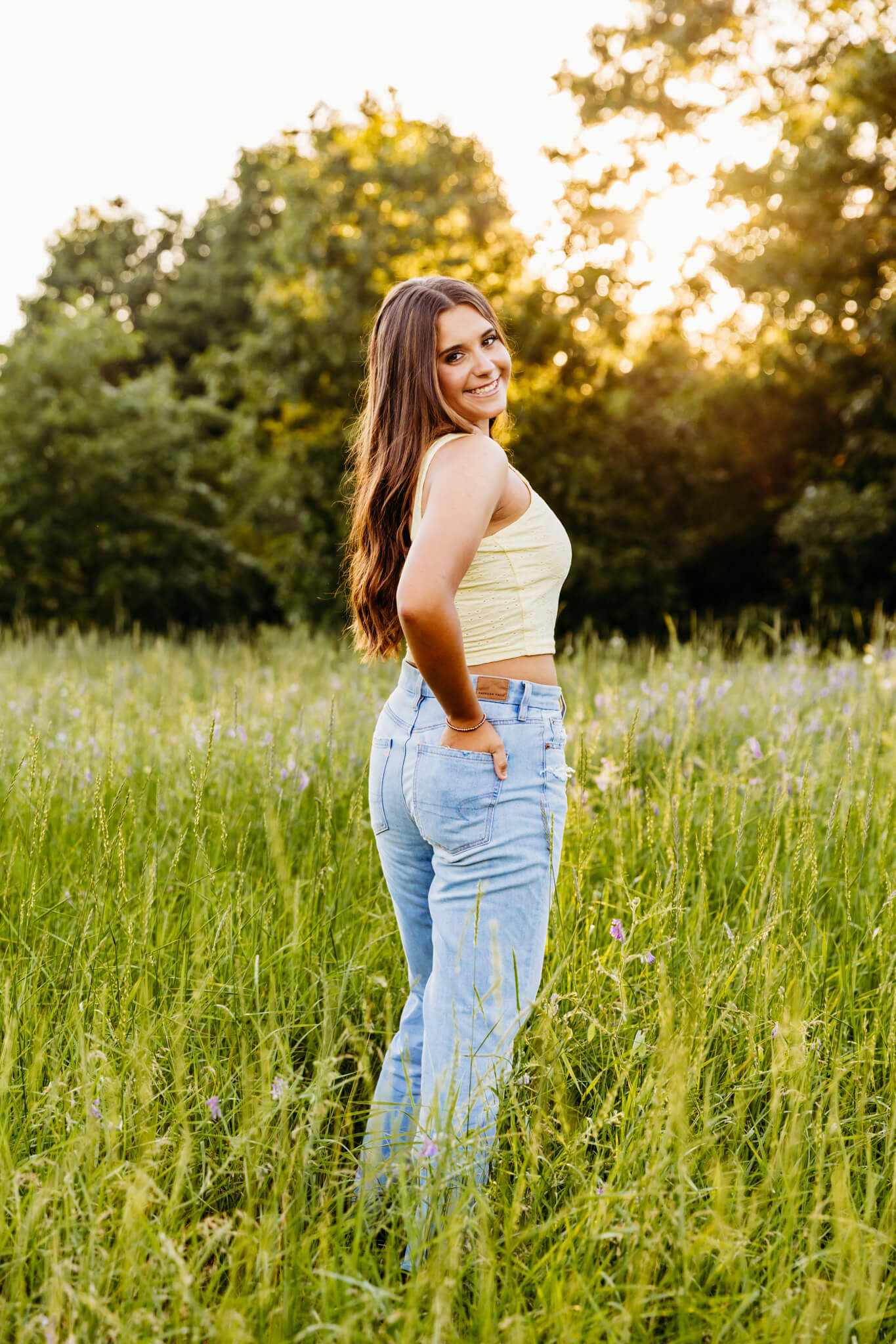 gorgeous student from Kimberly High School in jeans and a yellow tank top looking back and smiling as she stands in a wildflower field at sunset
