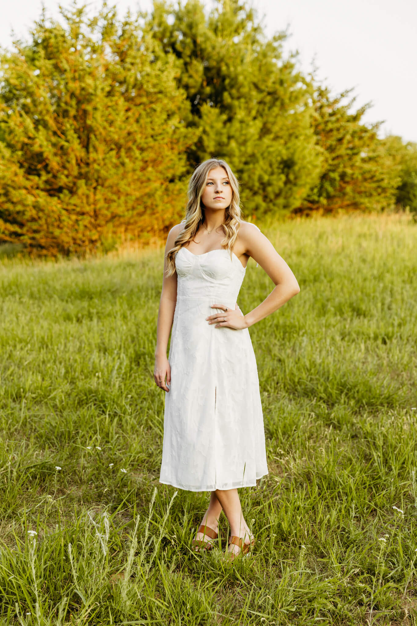 teen girl standing with hand on her hip in a white dress in a field at sunset