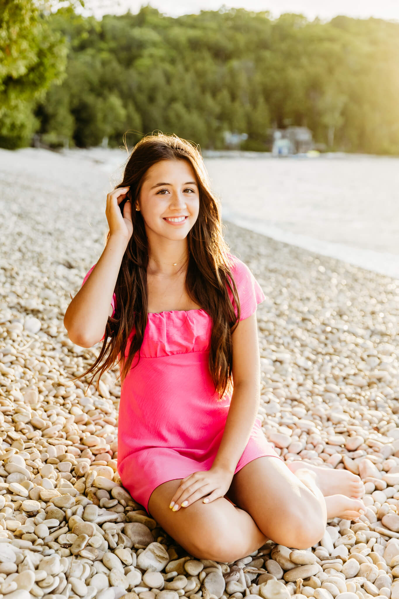 stunning teen girl in a bright pink dress tucking her hair behind her ears as she sits on a rocky beach for a blog post about Styled by Sharon