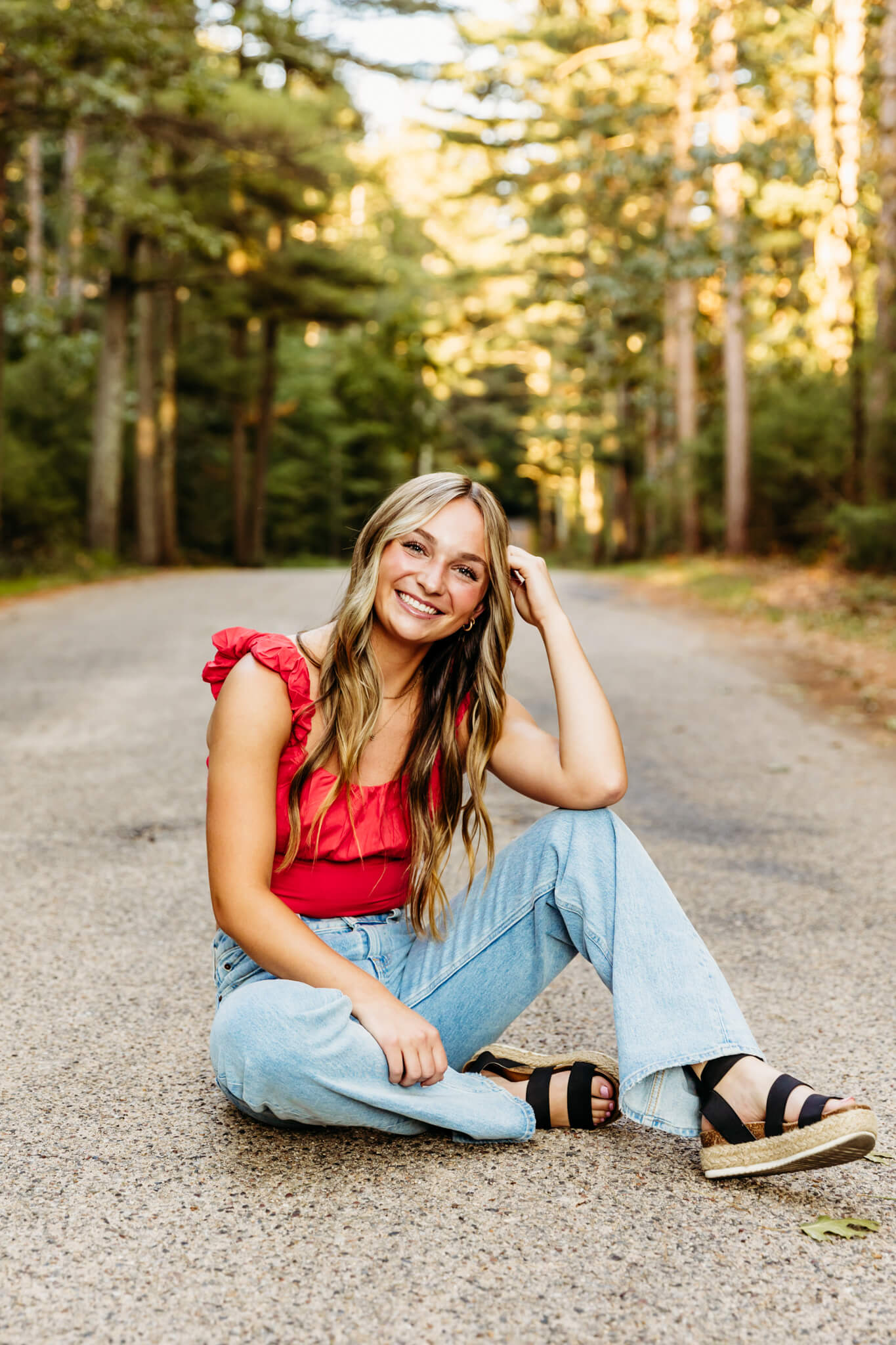 high school senior in a red top and denim bottoms sitting in the middle of a tree covered road for blog post about alterations in Green Bay