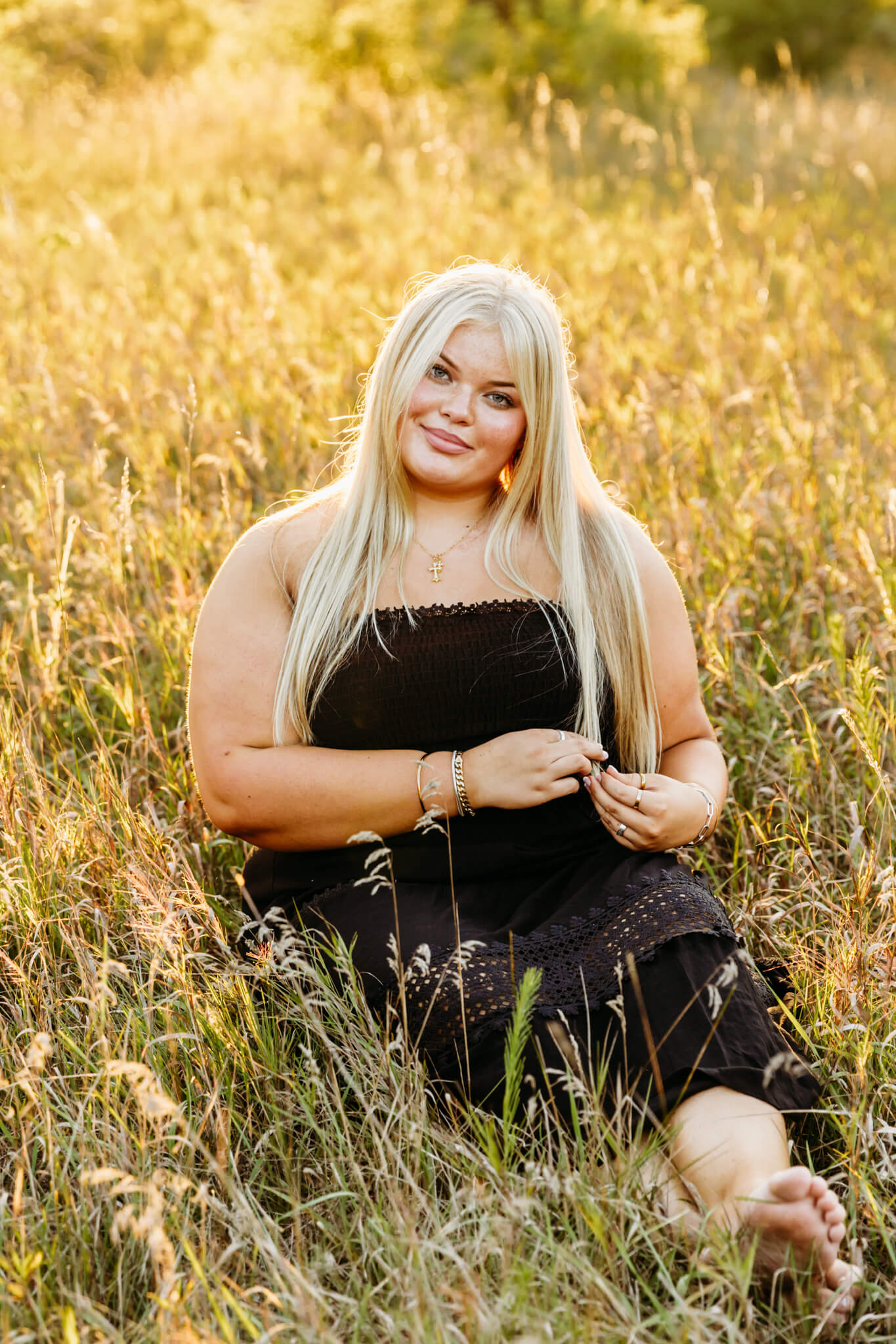 high school girl in a black dress sitting in a golden field playing with her hair for a blog post about Green Bay nail salons