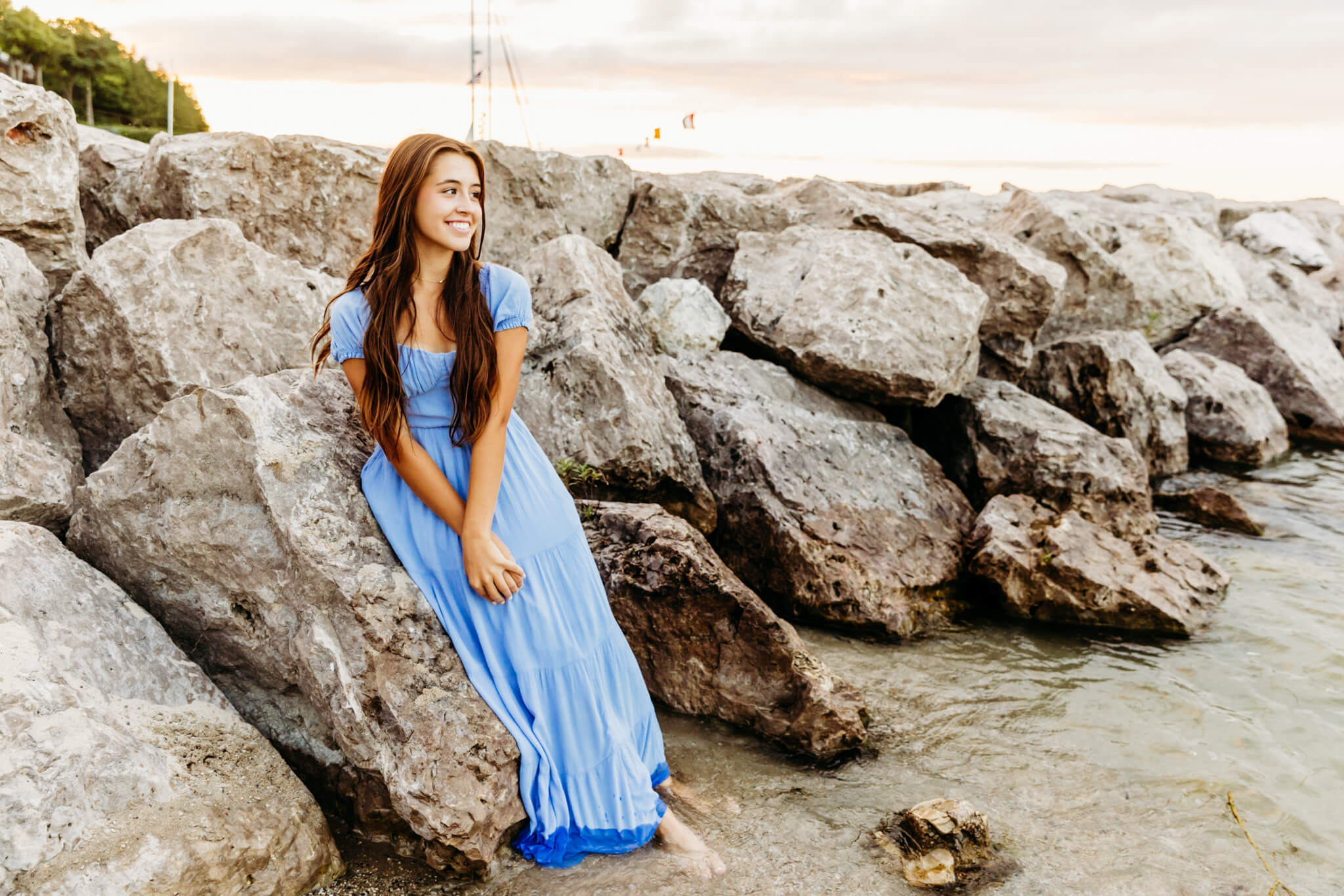 brunette teenage girl in a light blue dress sitting on a rocky shoreline with her feet in the water