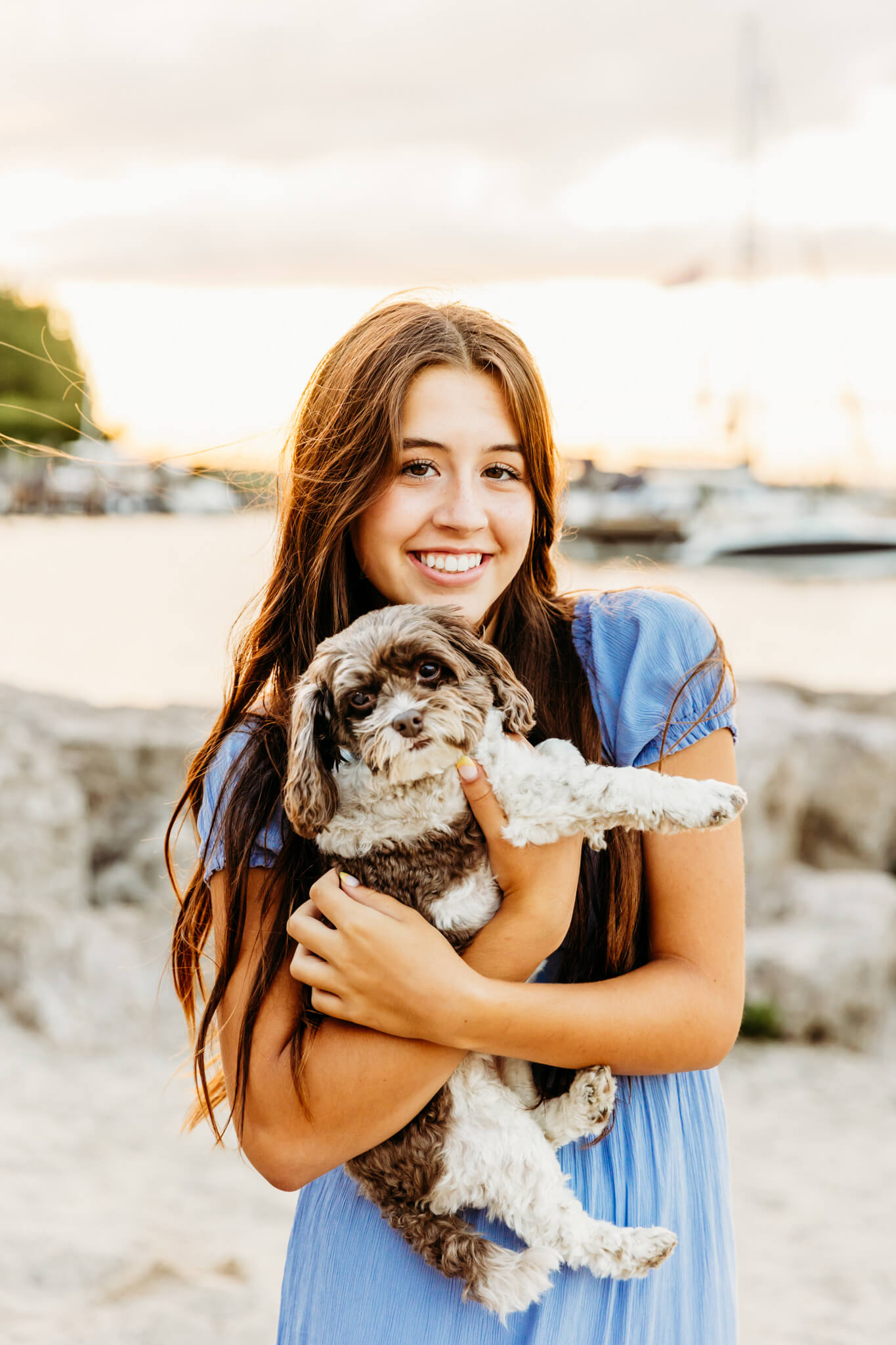 teenage girl in a blue dress snuggling her small dog on a beach at sunset near Appleton Wi