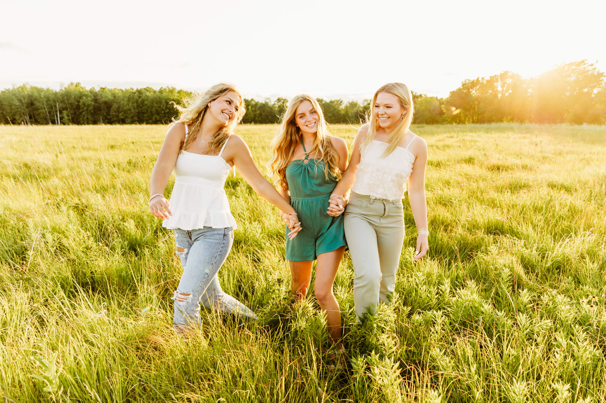 three high school senior girls holding hands while bumping hips in a grassy field near Oshkosh at sunset
