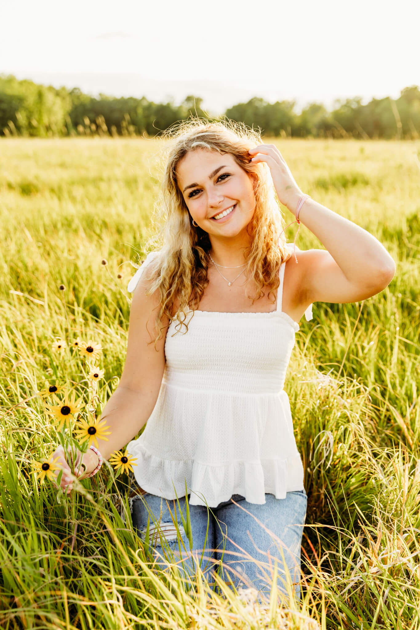 teen girl in a white top and jeans, pulling her hair behind her ears as she smiles and plays with the wildflowers for a blog post about tanning in Oshkosh WI