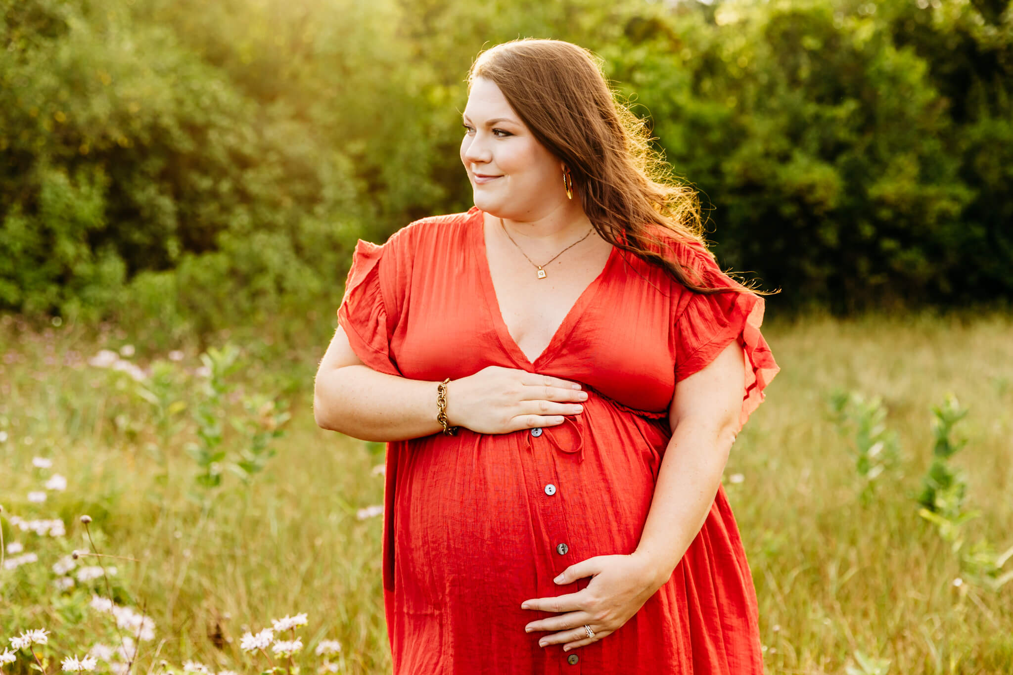 beautiful brunette mama to be in a bright orange dress holding her baby bump and glancing out over the field