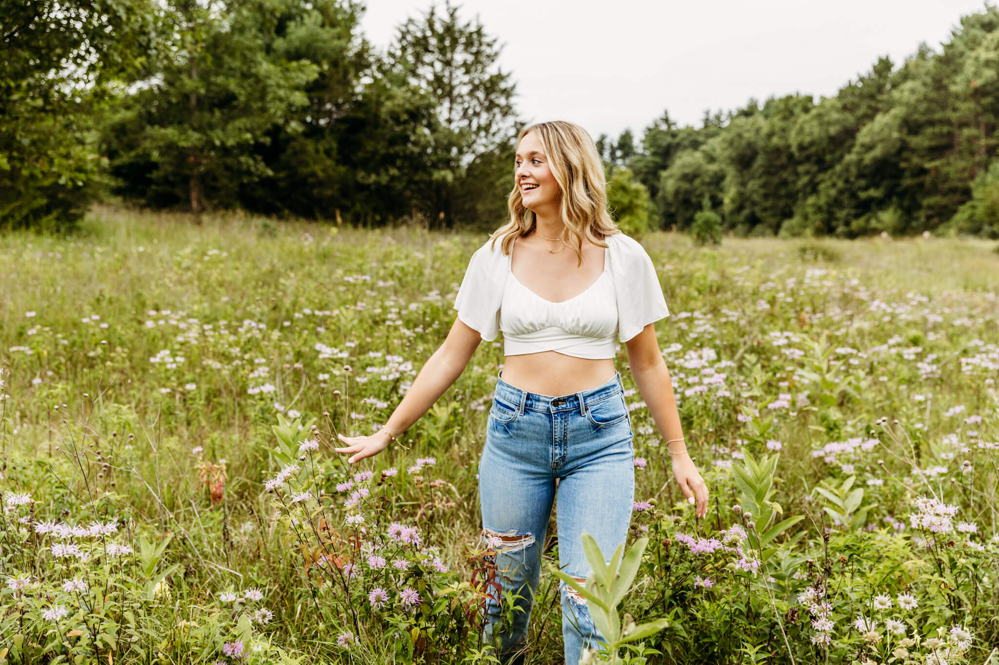 beautiful high school senior in a white crop top and jeans walking through a wildflower field near Oshkosh