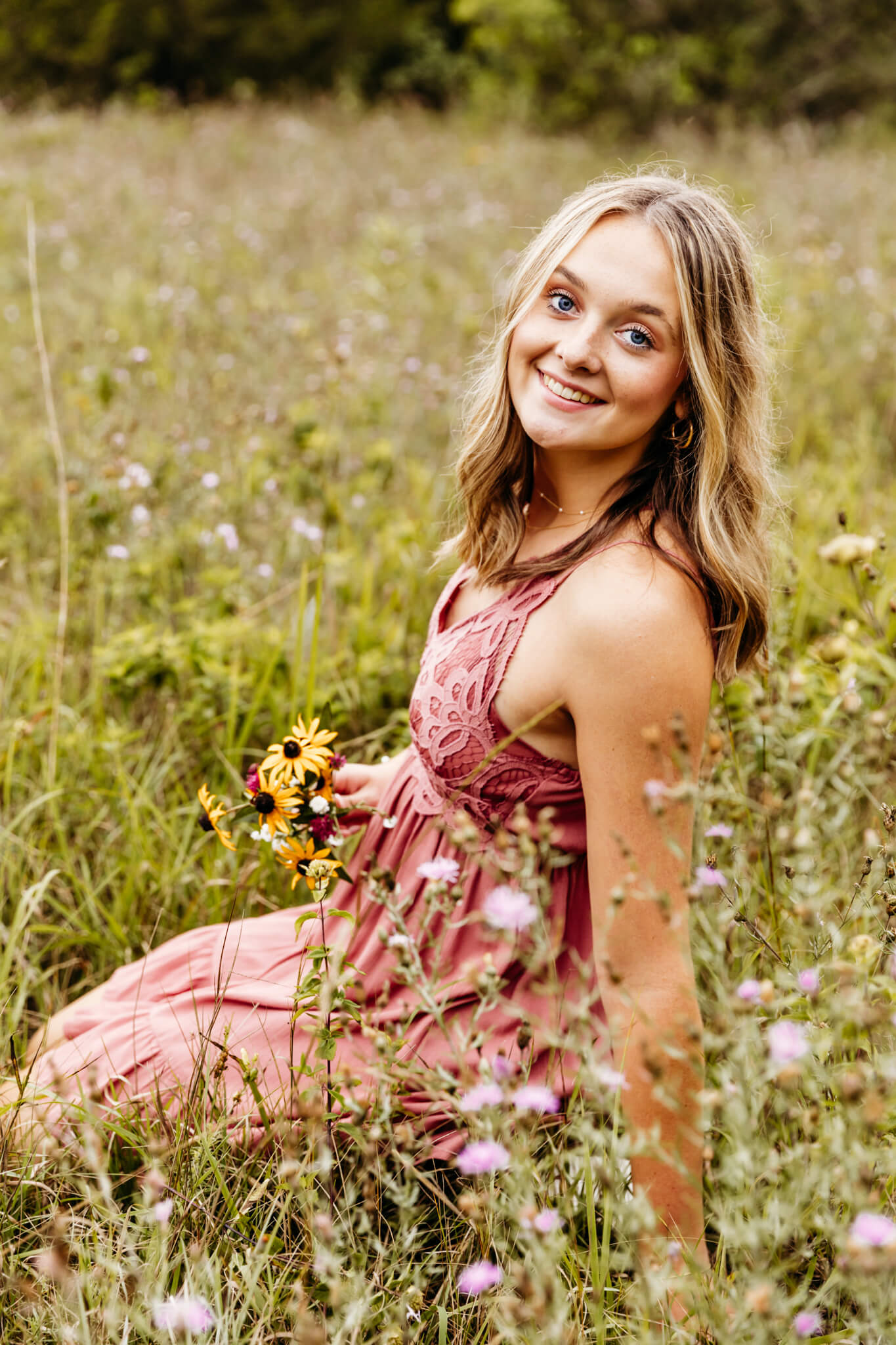 little chute high school senior in a pink dress kneeling in a field of flowers and smiling as she looks over her shoulder 