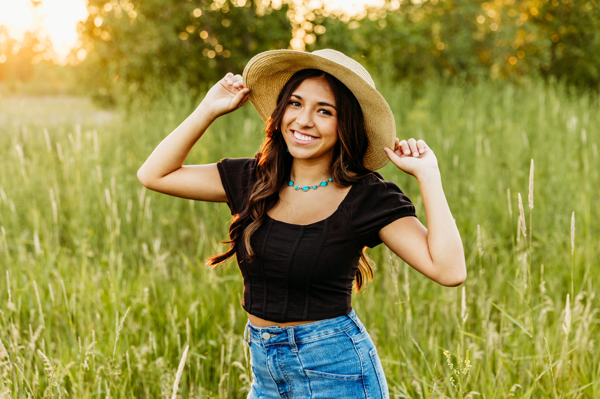young lady in a black top and denim pants standing with a straw sunhat on and her hands holding each side of the hat as the sun sets behind her