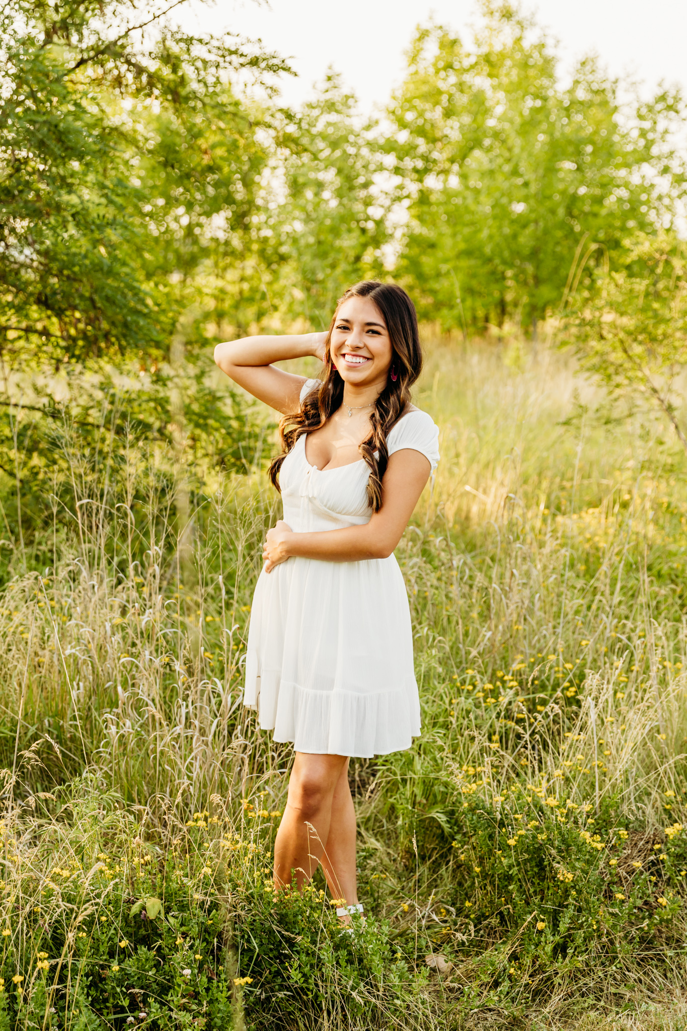 teenage girl in a short white dress standing in a field with a hand in her hair and the other across her body as she laughs