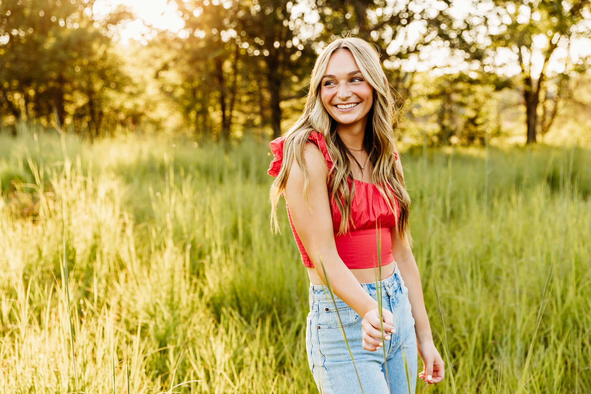 teen girl in a red crop top and jeans playing with tall grass and she smiles and looks over her shoulder in a glowing field at sunset near Green Bay