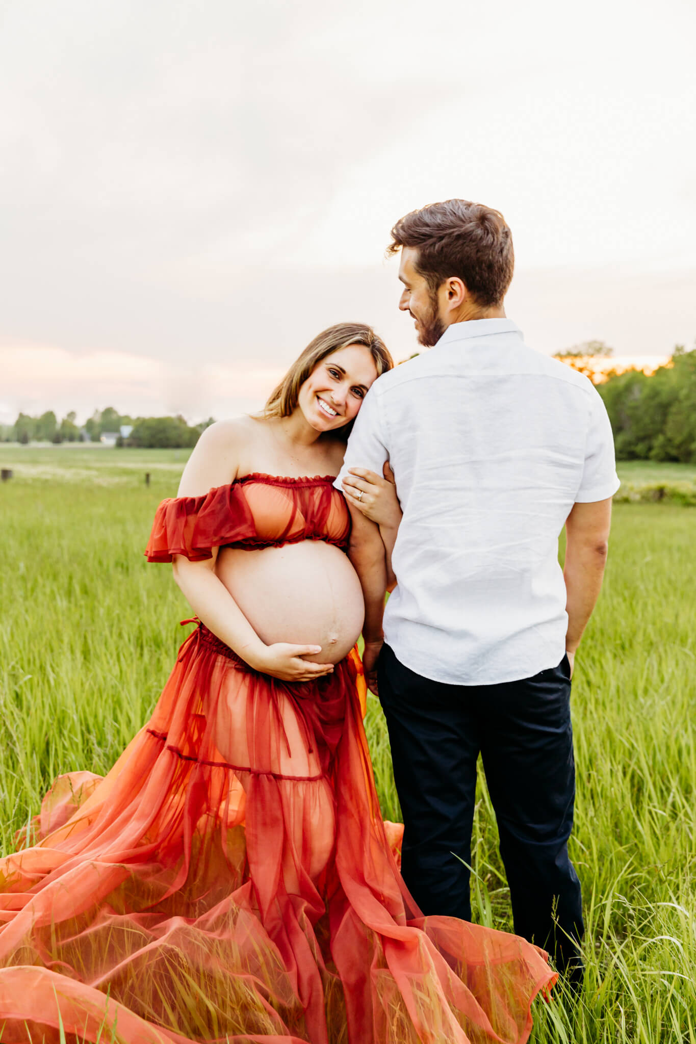 expecting mama in a two piece orange dress hugging her husbands arm and holding her baby bump for BirthWise Wellness in Appleton WI