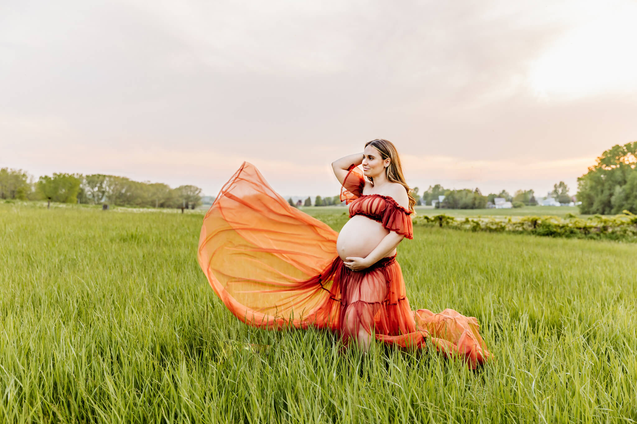 stunning pregnant mother throwing her dress in the wind as she gazes out into the field with the sunsetting behind her