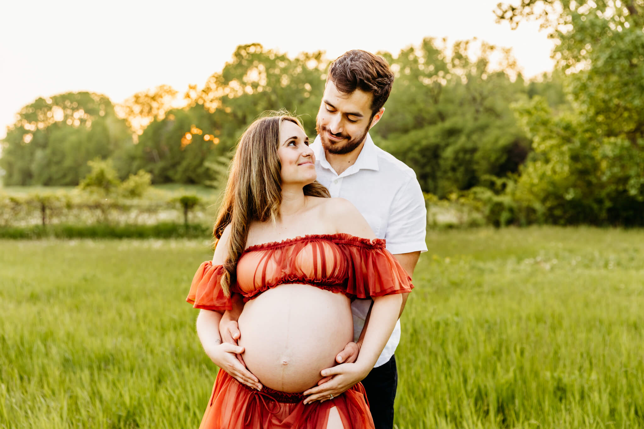 husband holding his wife from behind as they embrace their baby bump and smile at each other