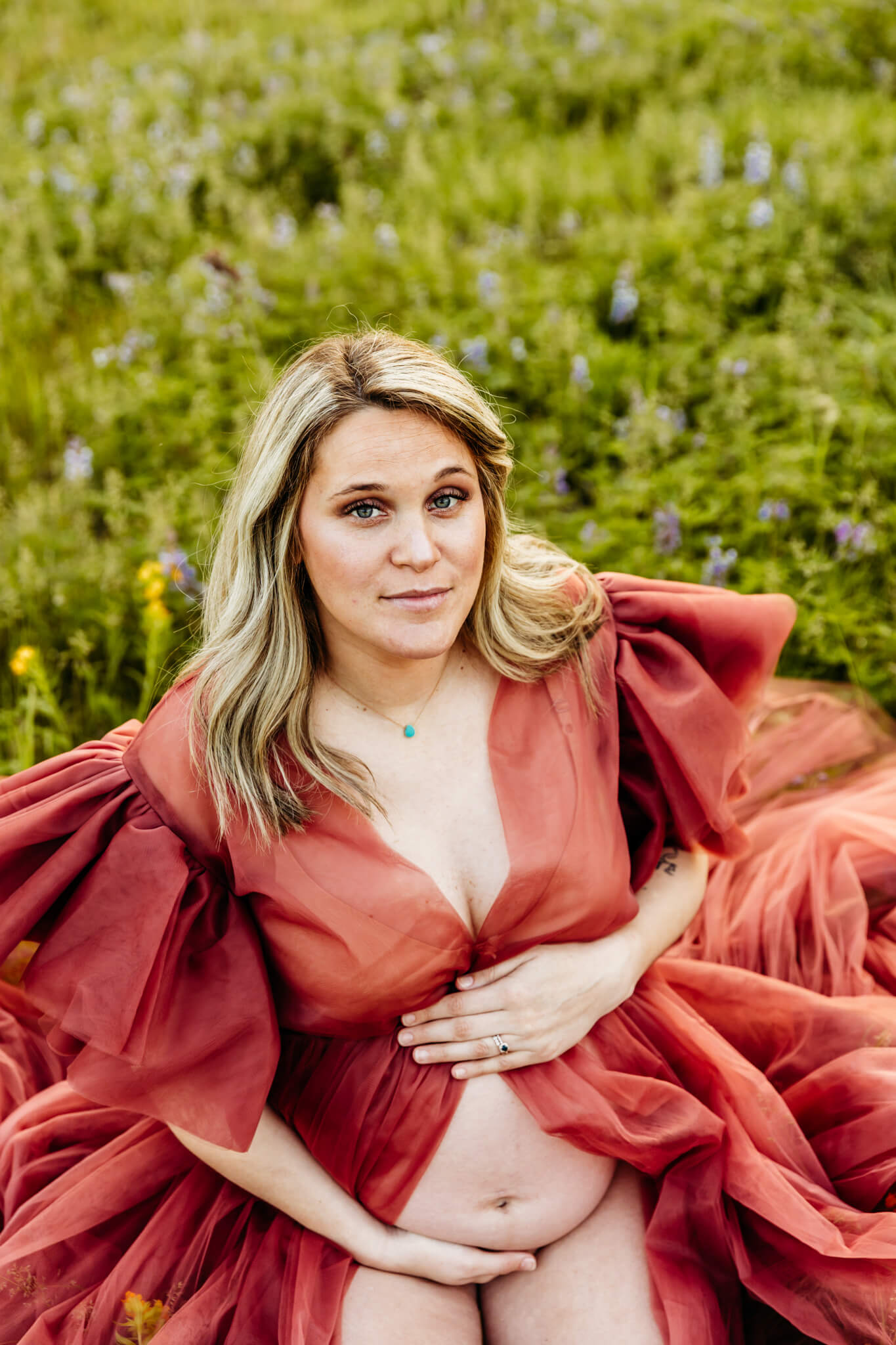 Mom to be, kneeling in a grassy field while holding her baby bump that's showing through her maternity gown as she glances up