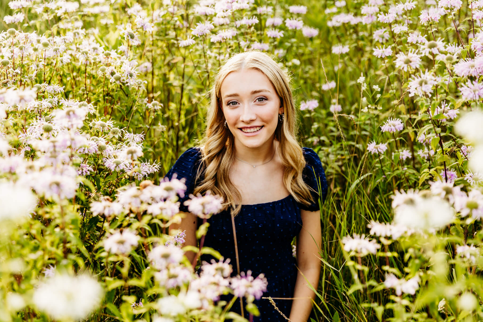 teen girl with blonde hair in a navy blue dress sitting in a field and is surrounded by purple wildflowers