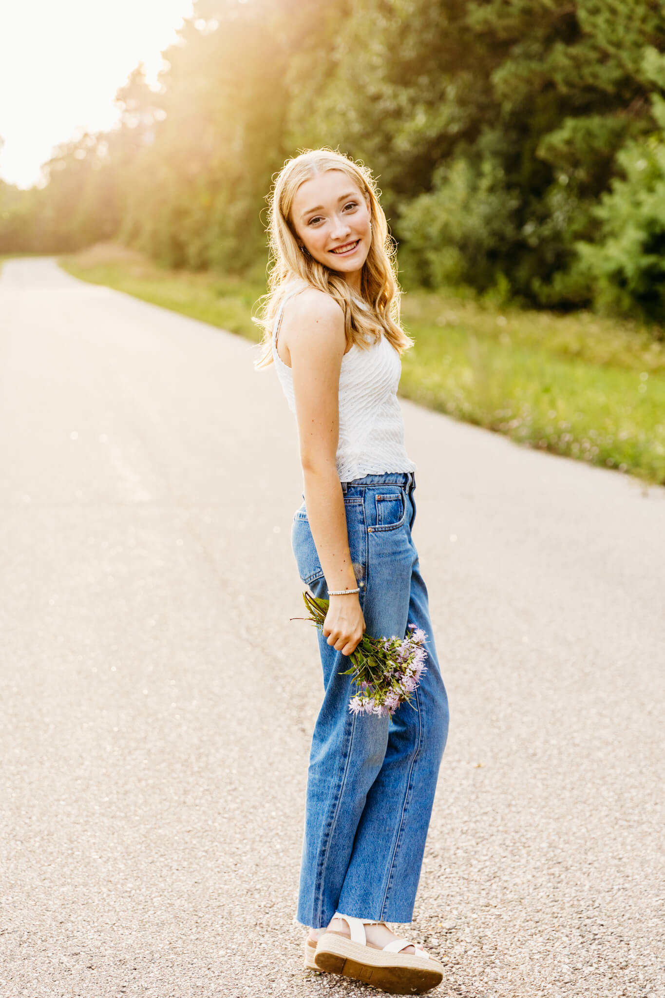 high school girl standing on a backroad with a bouquet of flowers at sunset