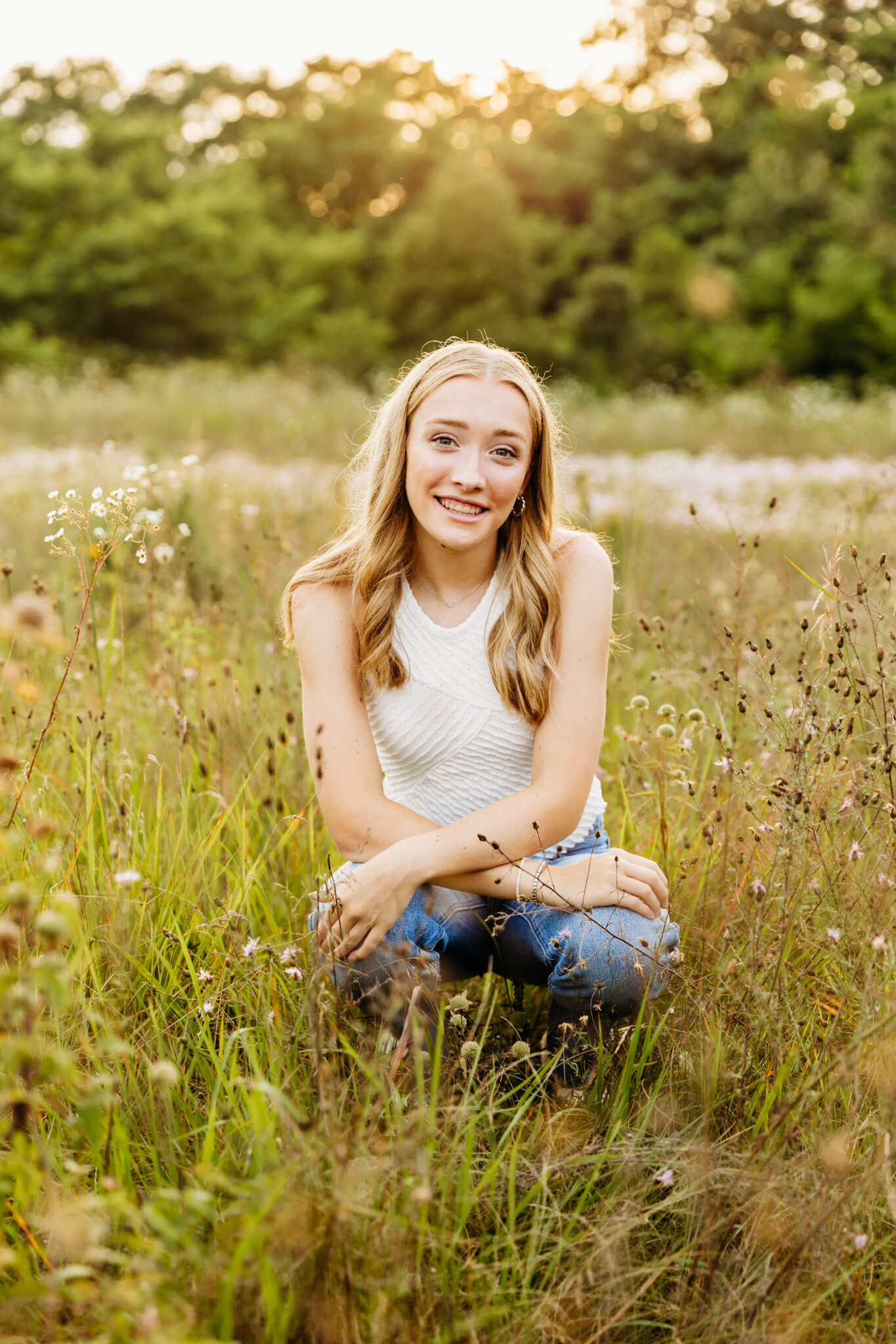 gorgeous high school senior with makeup by Aesthetics Layne kneeling in a field of flowers at sunset