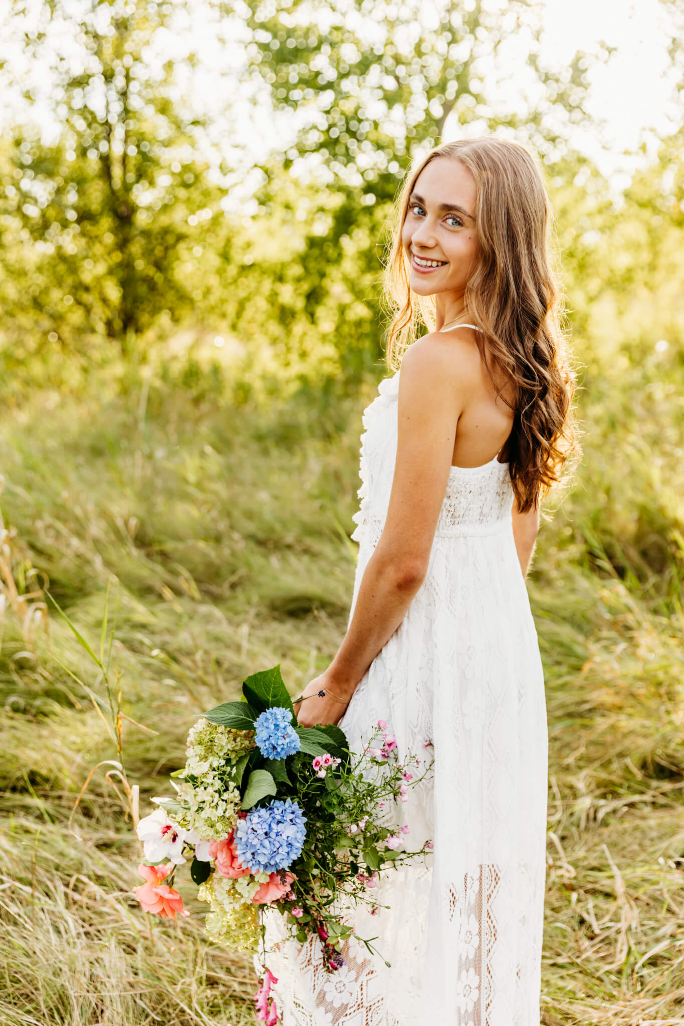 brown haired teen girl holding a flower bouquet by her side and looking back over her shoulder in a field at sunset.