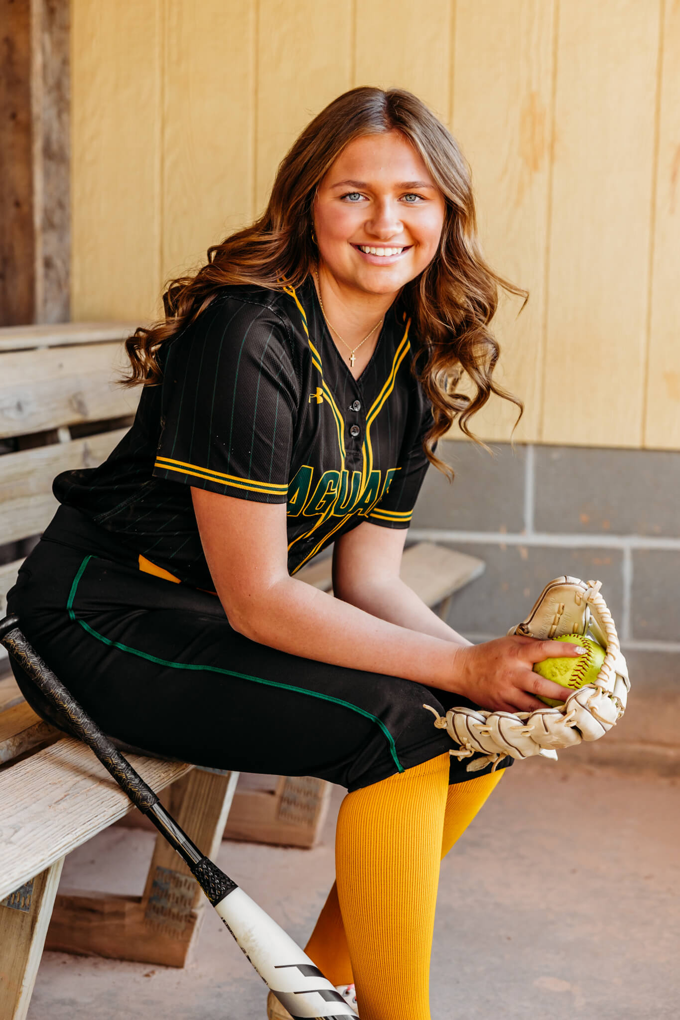 high school senior girl sitting on a bench in a dugout holding a softball in her glove