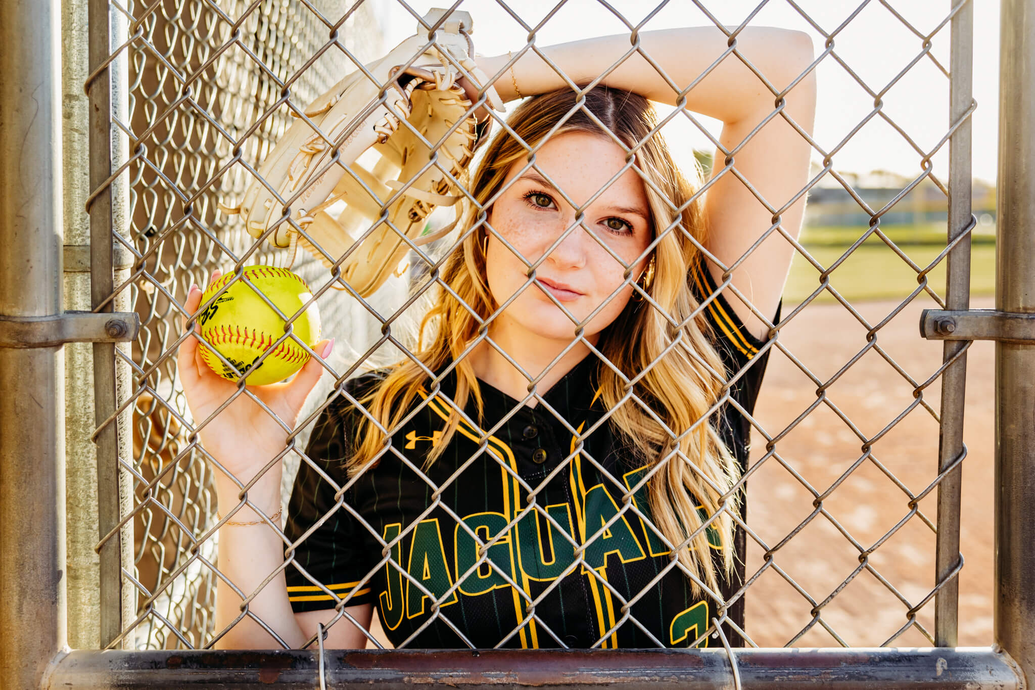 high school girl looking through a chainlink fence as she holds a softball and rests her wrist on her head while wearing her softball glove