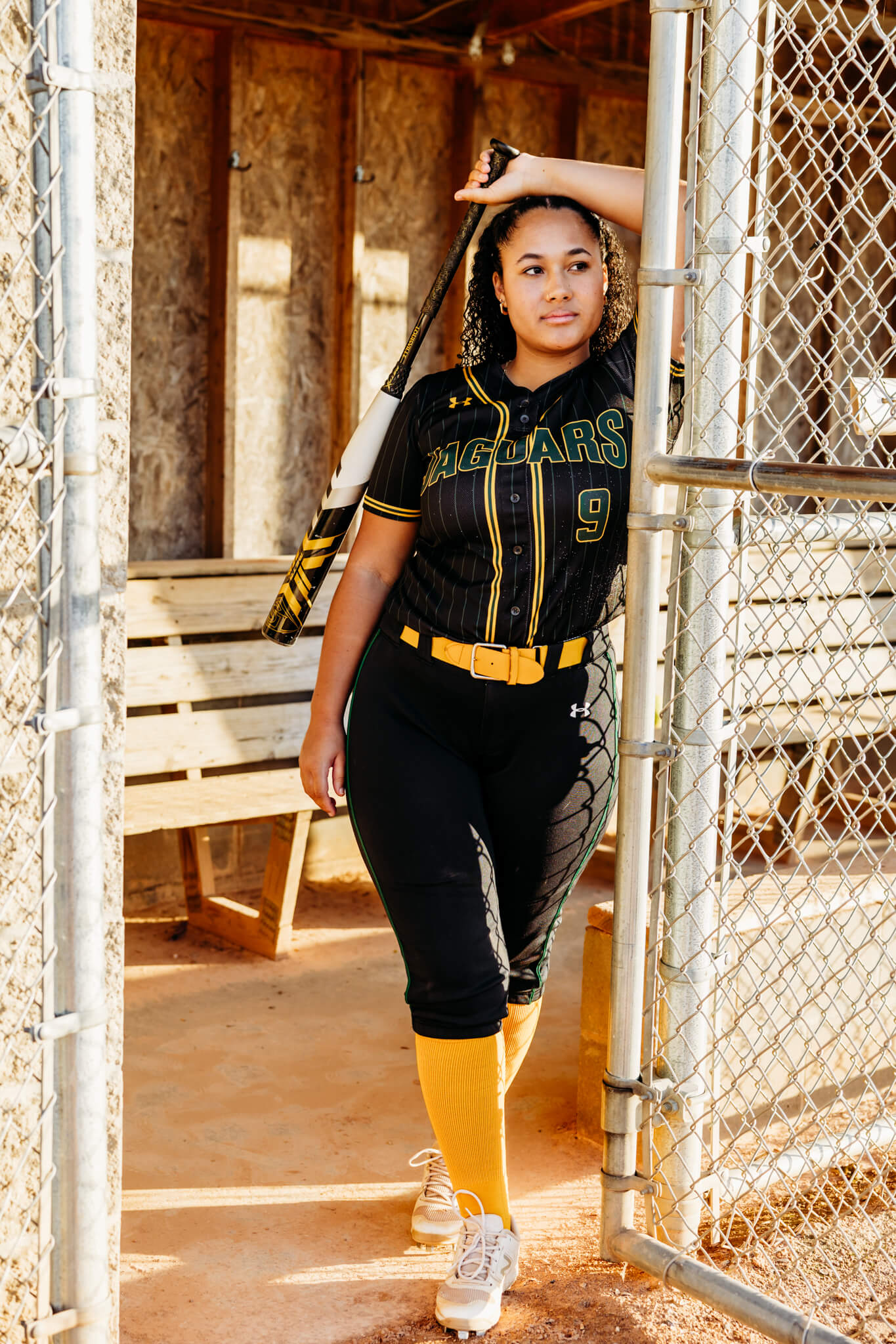 young lady in a softball uniform leaning against the door into a dugout and resting her wrist on her head as she holds a softball bat and looks into the field for senior photos