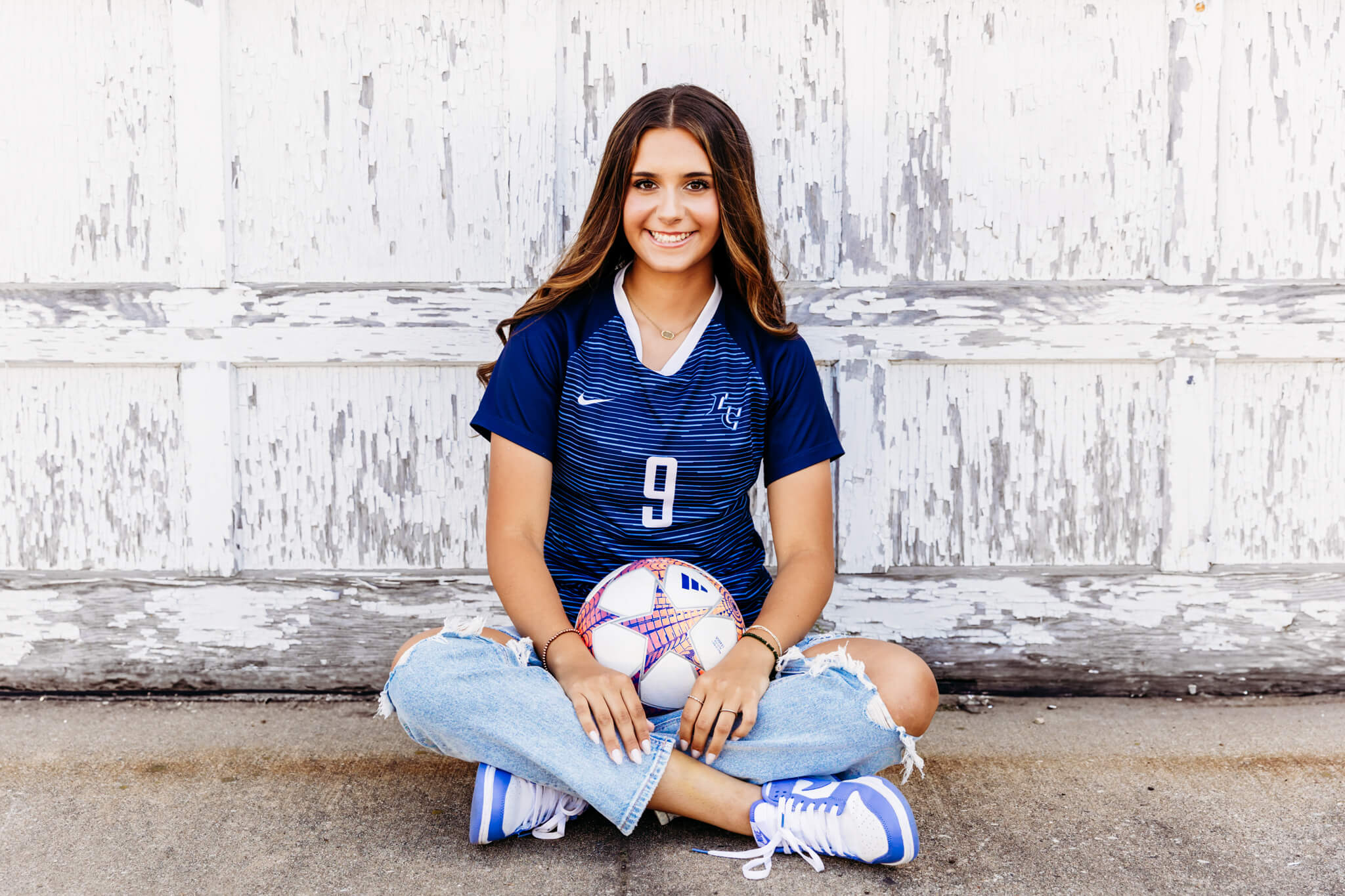 high school student athlete sitting in front of a rustic, white garage door and holding her soccer ball as she smiles proudly