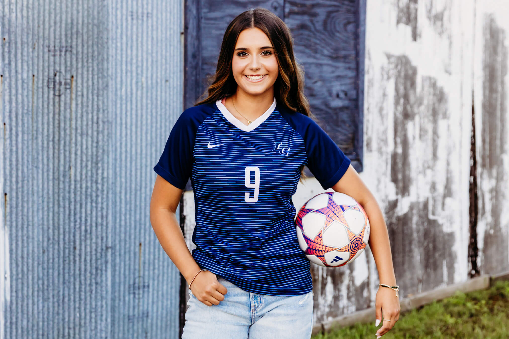 high school girl smiling as she holds her soccer ball on her hip while wearing her soccer jersey and jeans near downtown De Pere.