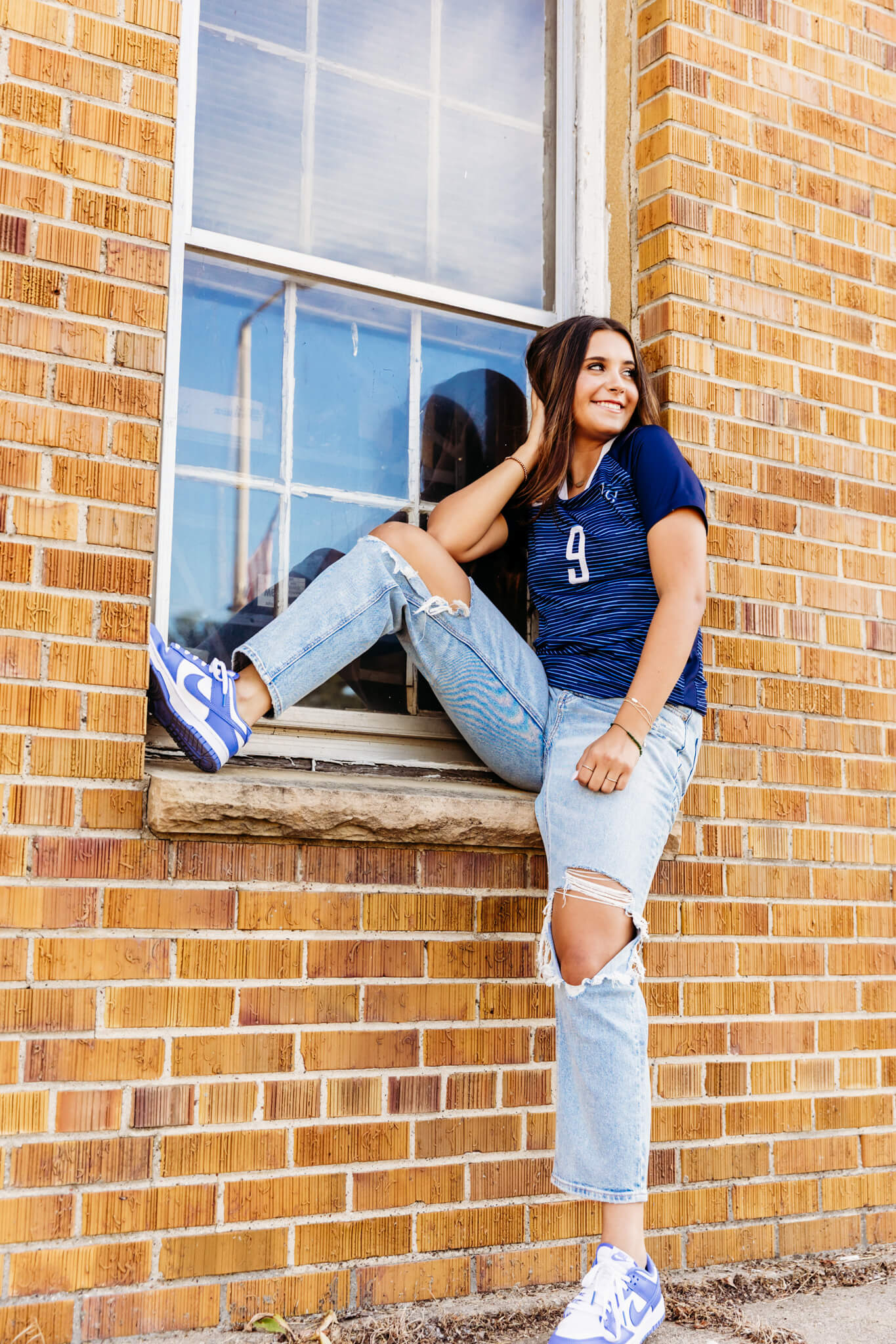 teenage girl in her soccer jersey sitting on a ledge by a window in a brick building