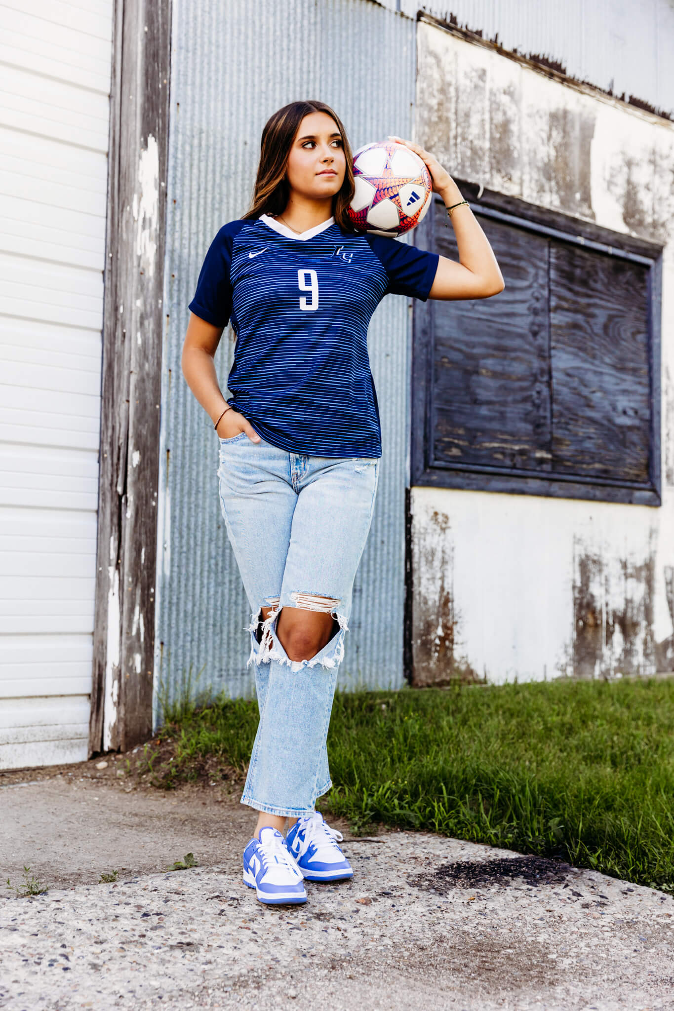 high school girl holding a soccer ball on her shoulders in front of a rustic building