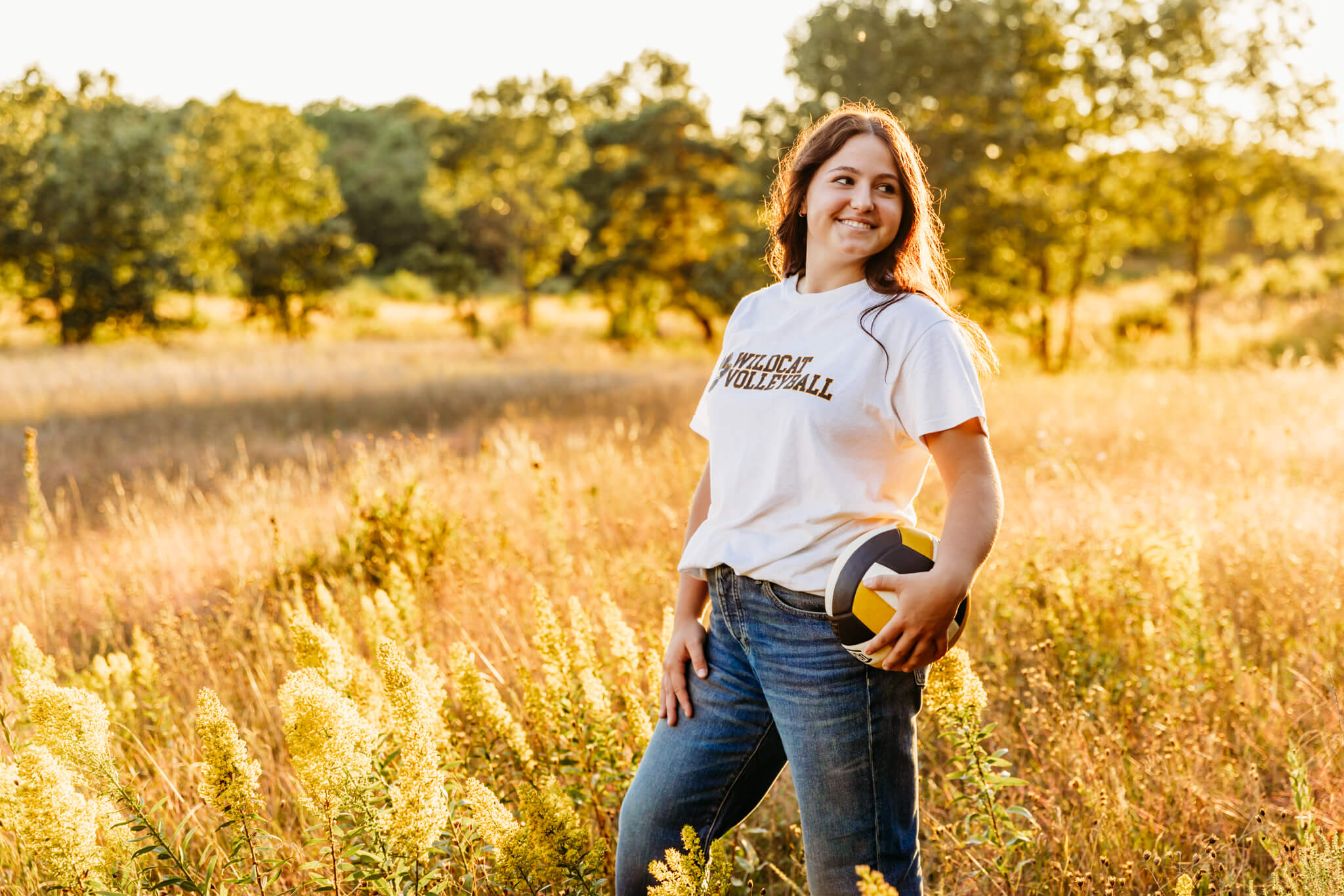 high school girl standing in a glowing field at sunset while holding a volleyball at her hip. 