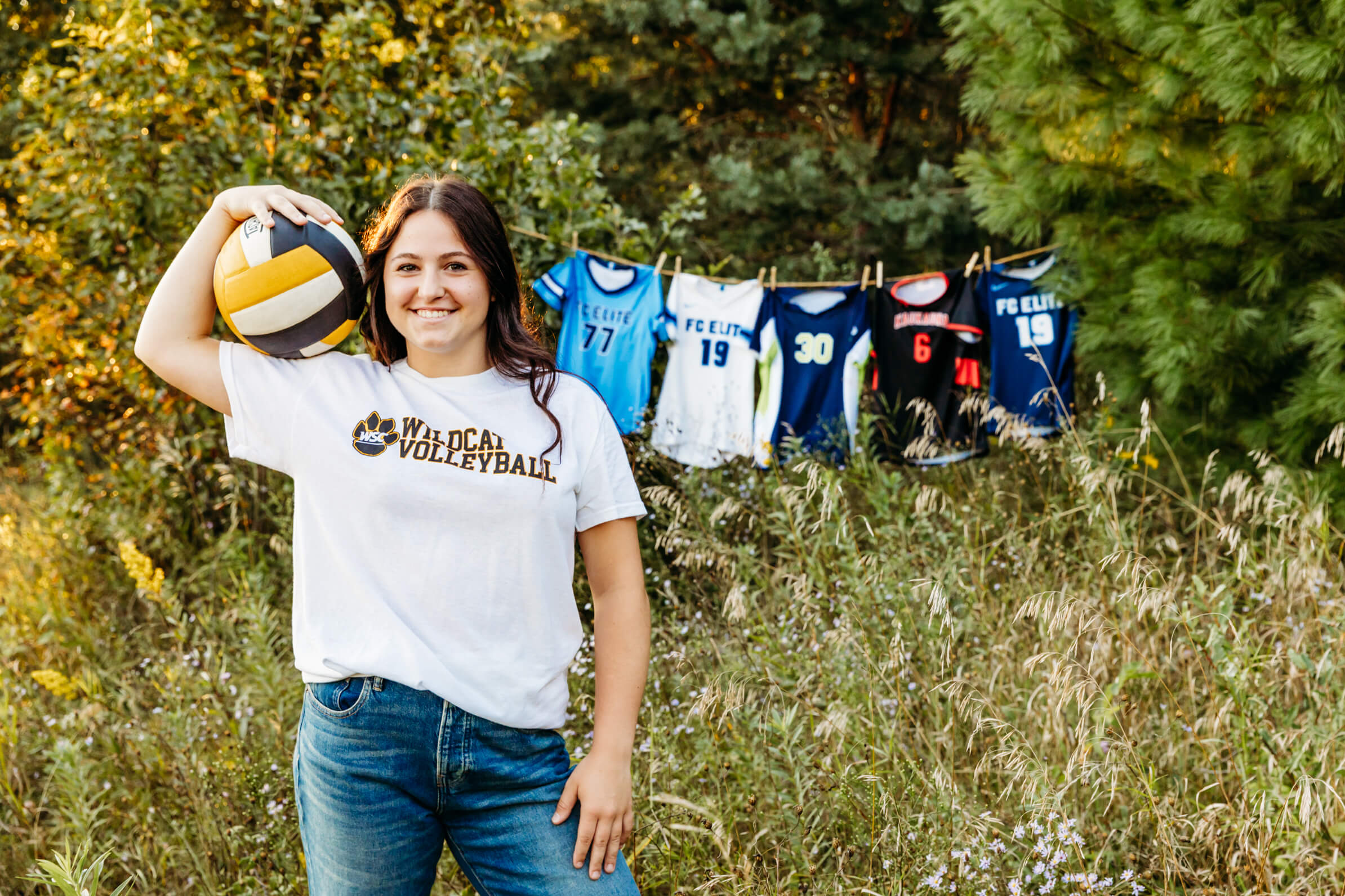 teen girl holding a volleyball on her shoulder while standing in front of her volleyball jerseys for her senior photos.